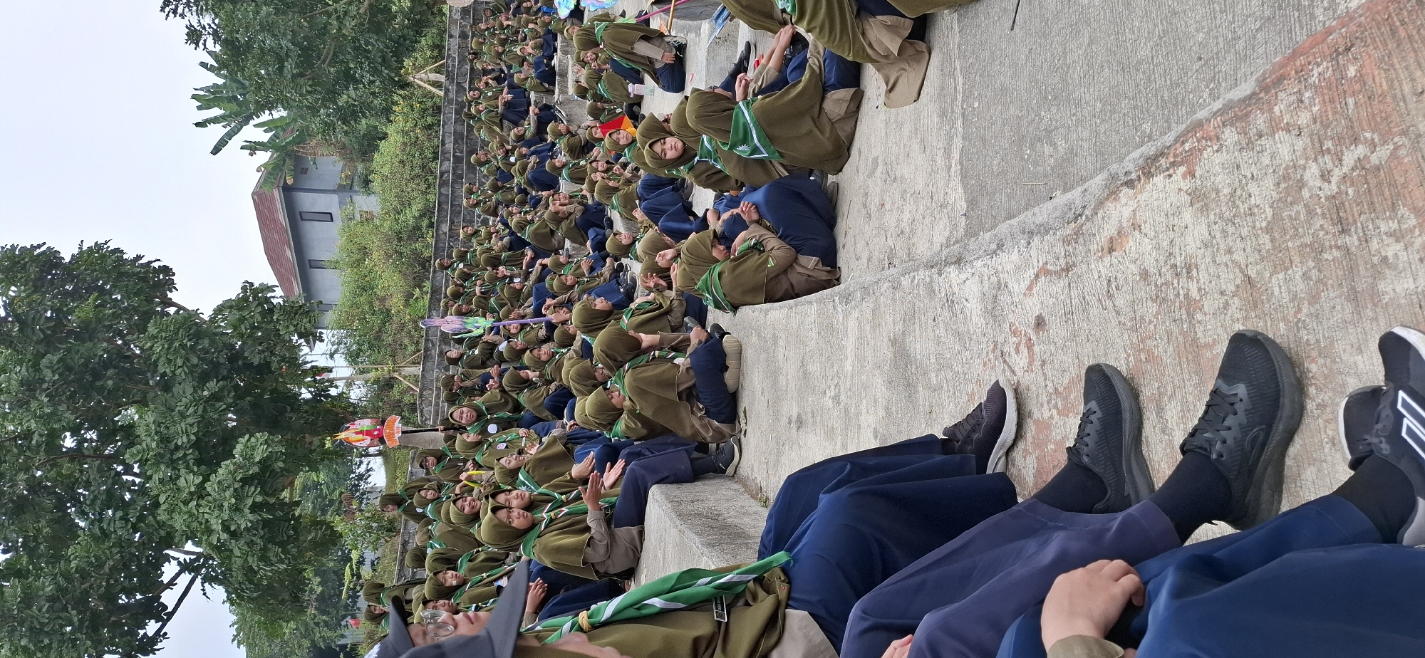 A group of young girls, dressed in khaki scout uniforms with green scarves, are seated on concrete bleachers. The scene appears to be outdoors, possibly in a park or an open-air venue, with trees and a building visible in the background. The girls are facing towards the left of the frame, with some looking towards the camera. One girl in the foreground, on the far left, is wearing a black baseball cap and is looking to her left. The bleachers are tiered, and the girls are arranged in rows, suggesting a gathering or event. The overall atmosphere seems to be casual and relaxed. The location is identified as Karang, Indonesia.