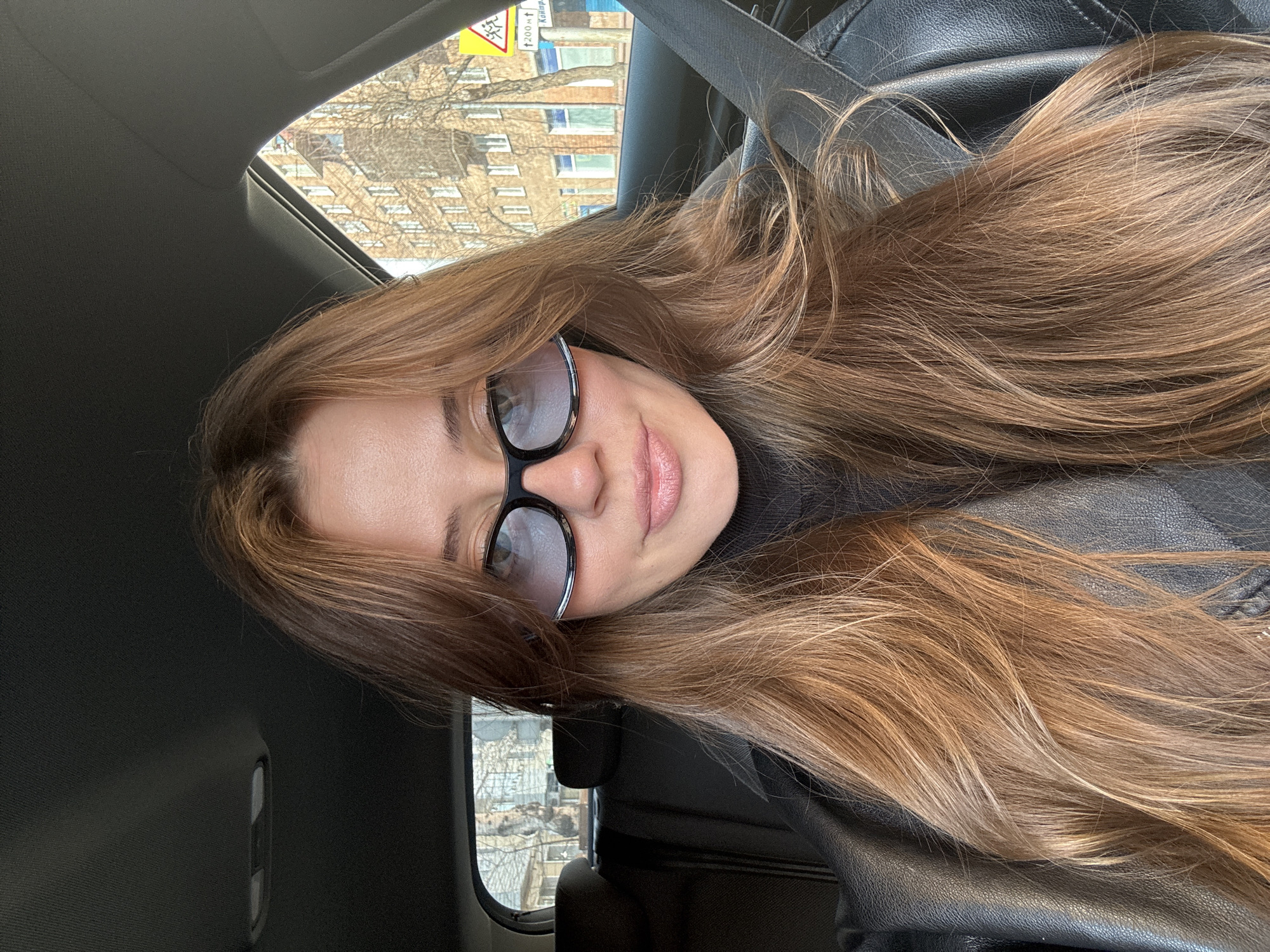 A woman with long, light brown hair is in a car. She is wearing sunglasses and a dark top. The background visible through the car window shows a building, suggesting an urban environment. A road sign is visible in the top of the window, with 200 meters to Kiev street, indicating the location as Krasnoyarsk, Russia.