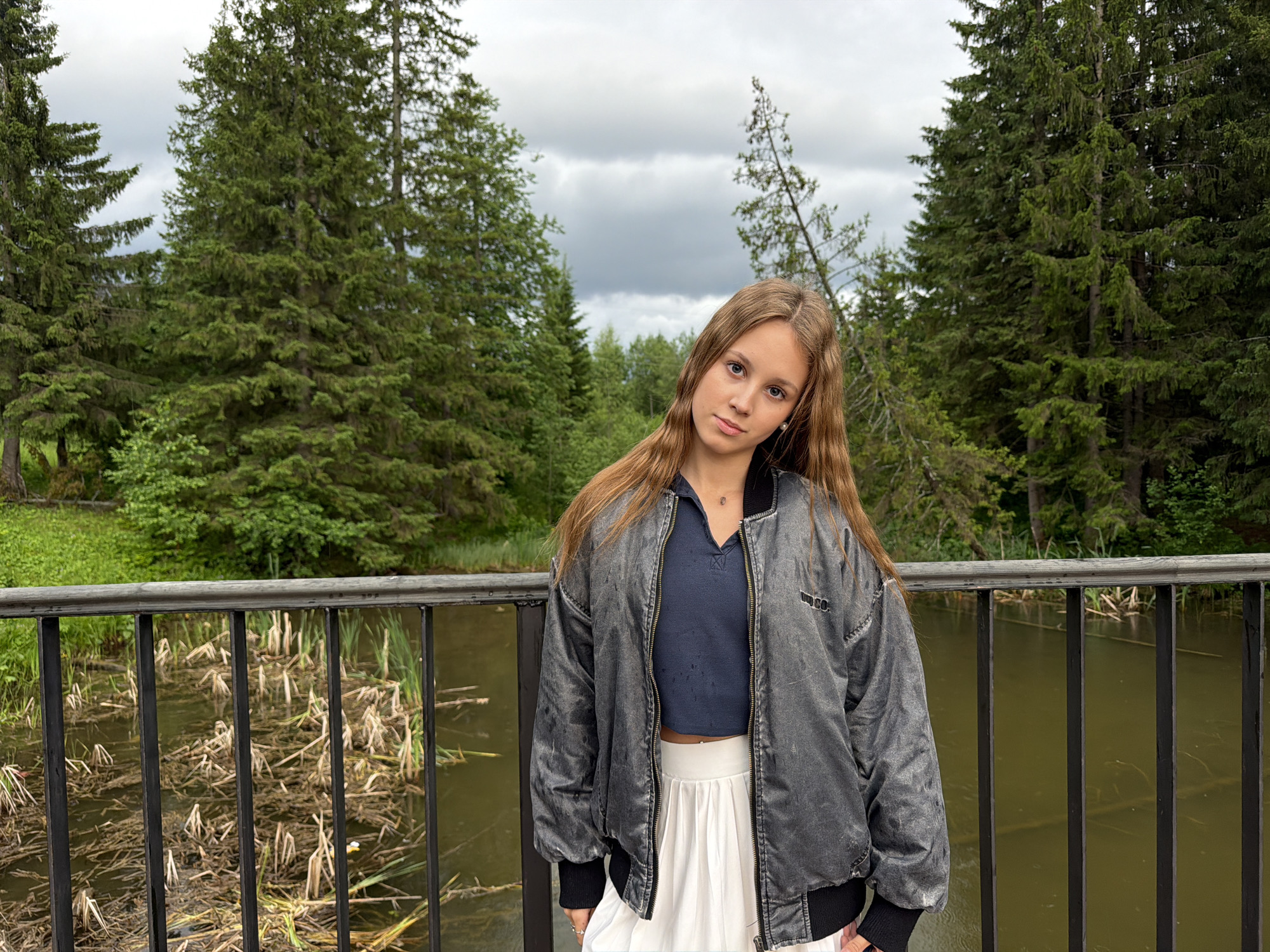 A young woman stands outdoors on a cloudy day, leaning slightly against a black metal railing. She has long, light brown hair and is looking at the camera. She is wearing a dark blue V-neck crop top, a dark gray bomber jacket with a zipper, and a white pleated skirt. The background features a body of water with reeds and fallen branches, bordered by a lush green forest of tall pine trees. The overall setting appears to be natural and possibly a park or recreational area, consistent with locations like those found in Chusovoy, Russia.