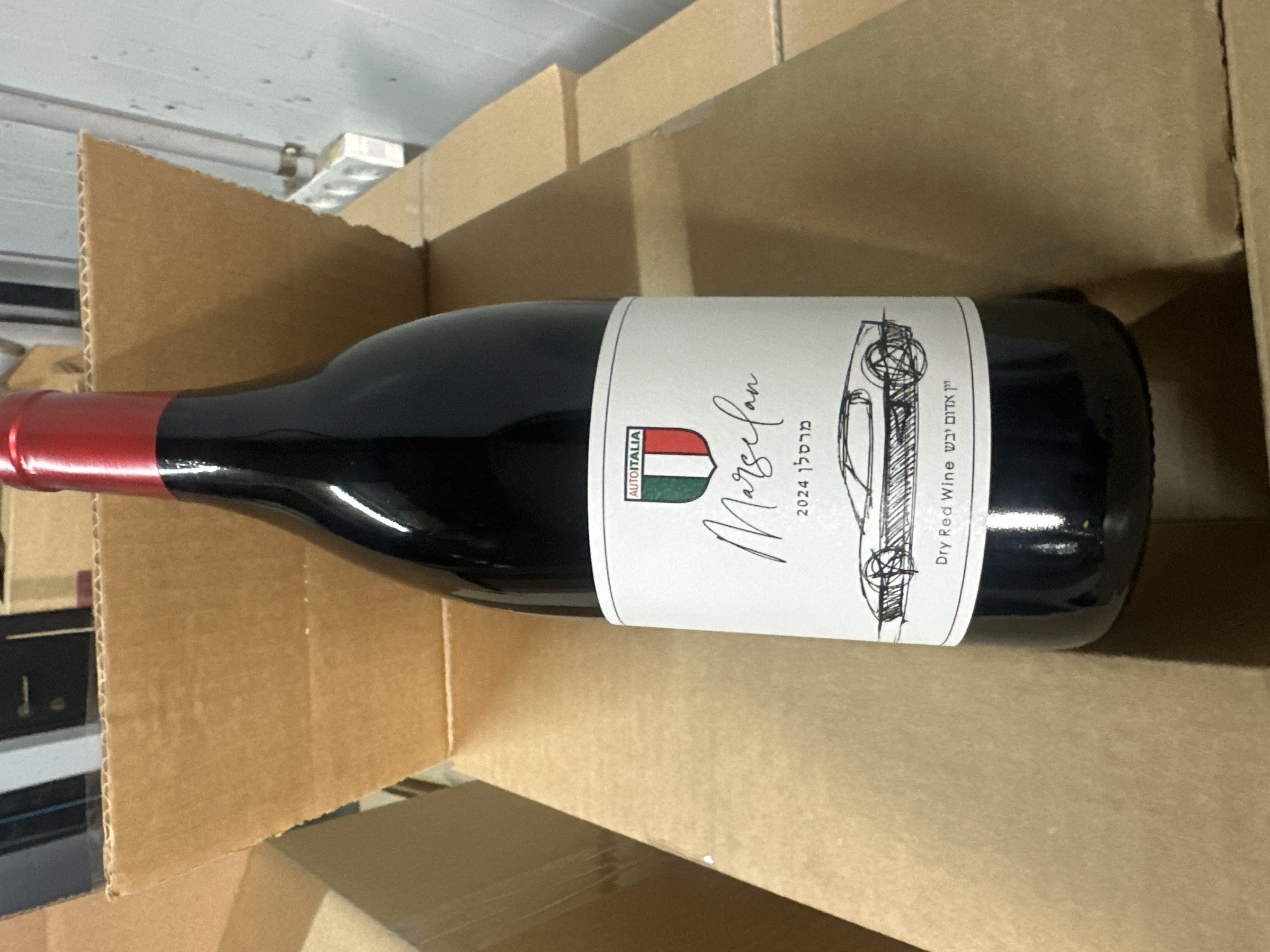 A dark glass wine bottle, sealed with a red foil capsule, stands vertically within an open, light brown corrugated cardboard box. The bottle's white label displays a shield logo with green, white, and red vertical stripes, surmounted by the text "AUTOITALIA". Below the logo, "Marselan" is written in script, followed by "2024" and the Hebrew word "מרסלן". An outline drawing of a classic sports car is depicted beneath this text, with "Dry Red Wine" and the Hebrew words "יין אדום יבש" printed at the bottom of the label. The immediate environment consists of the interior of the cardboard box. In the background, additional stacked cardboard boxes are visible, along with a light-colored wall featuring a vertical pipe or conduit on the right. This scene is situated in an indoor storage or warehouse facility in Tira, Israel. No individuals are present in the image.