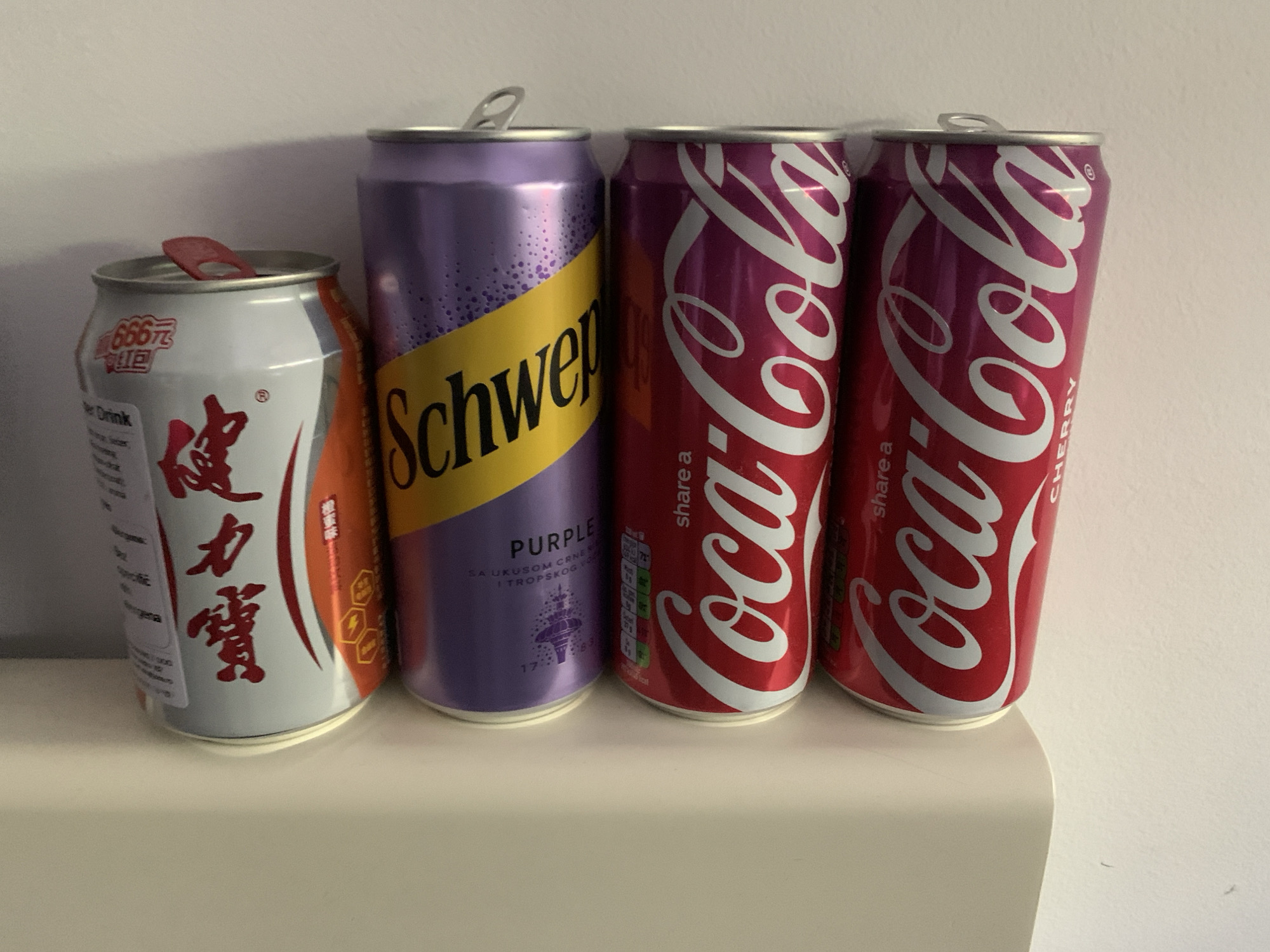 The image shows four cans of soda lined up on a white surface. From left to right, they are: an opened can with white and red labeling, a purple Schweppes can with a yellow diagonal stripe, a red Coca-Cola can, and a red Coca-Cola Cherry can. The backdrop is a plain white wall. Location data is not visible in the image.