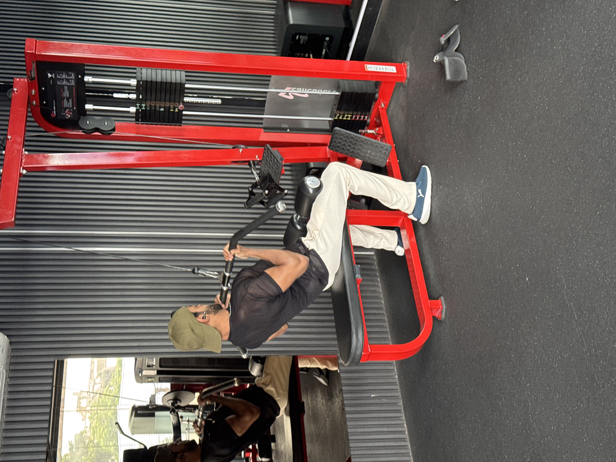 A man wearing a black t-shirt, beige pants, and a green cap is performing a lat pulldown exercise on a red weight machine in a gym. The gym has dark gray ribbed walls and a black rubber floor. A large mirror reflects the man and the gym equipment. In the background, a black machine with a digital display is visible, and the machine has a stack of black weights. The scene appears to be a modern gym. There is no indication that the location is in Kolkata, India.