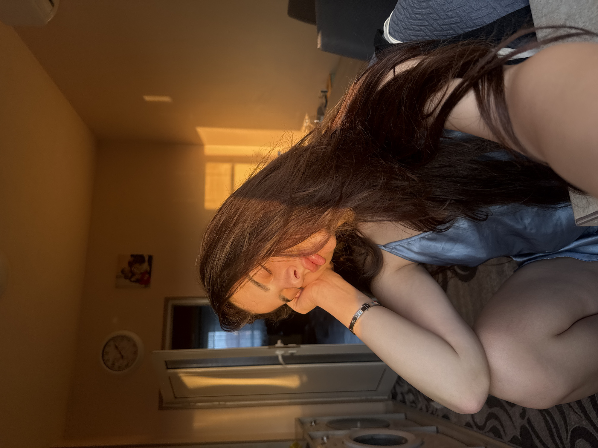 A selfie portrait depicts a young adult female seated indoors, her head resting on her right hand with eyes closed and lips slightly pursed. She has long, dark brown hair and wears a light blue satin camisole, with a black bracelet featuring a silver design on her right wrist. The scene is illuminated by warm, golden light, casting strong highlights and shadows, suggesting either sunrise or sunset.
The background shows a domestic interior with light brown walls. To the left of the subject, two white front-loading appliances, possibly washing machines, are visible next to a partially open white door that reveals a darker room with a window covered by horizontal blinds. Above the door, a white circular wall clock displays approximately 10:15. Further right on the wall, a small, indistinct framed picture is visible. To the right of the subject, bright rectangular patterns of sunlight are cast on the wall, and a portion of a dark grey upholstered chair or sofa is present. The floor is covered by a patterned rug with grey and white geometric designs. An air conditioning unit is partially visible in the top right corner of the room. The visual content of the image does not provide any specific indicators to forensically confirm the location as Plovdiv, Bulgaria.