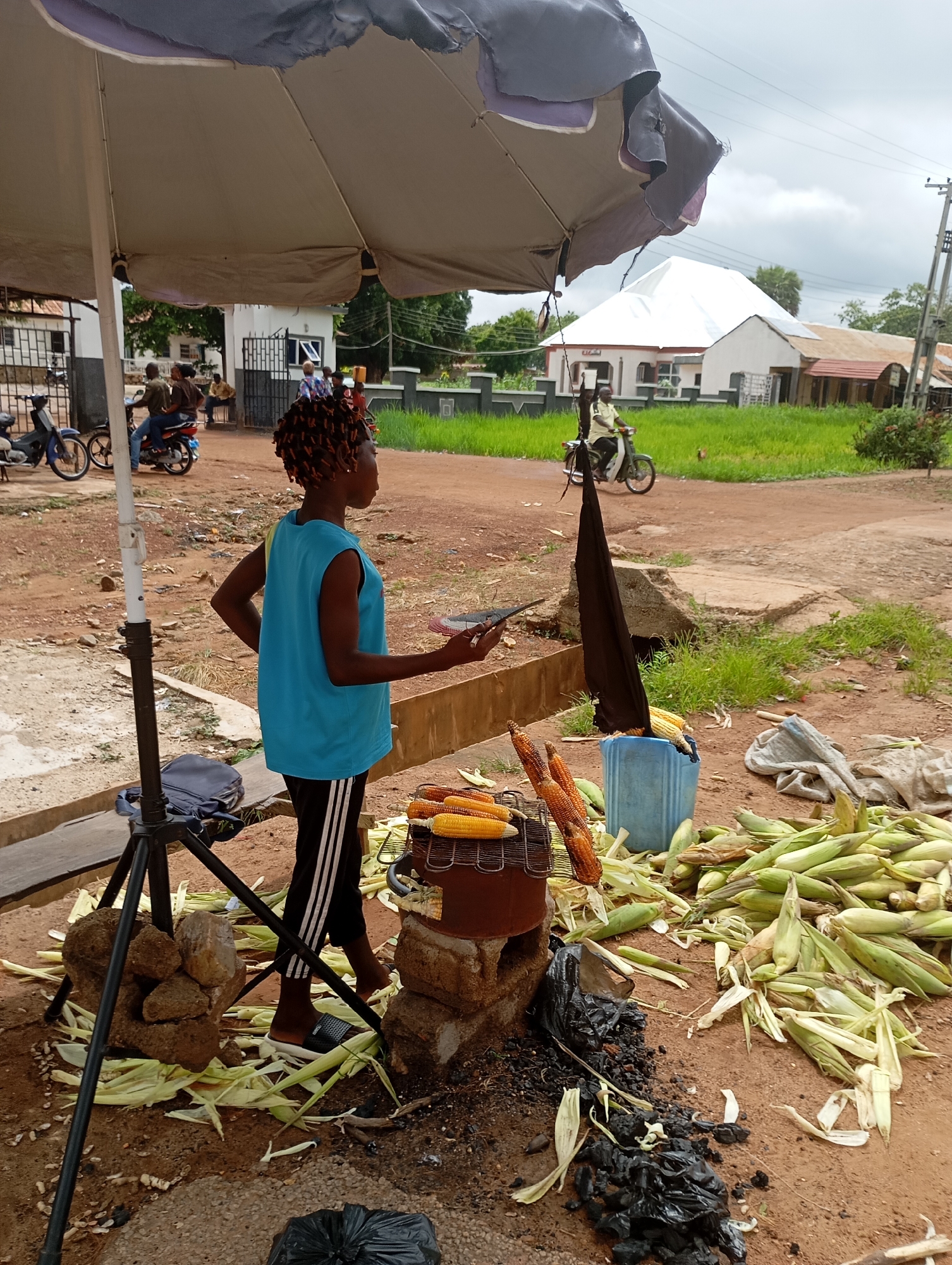 A young female vendor stands under a large umbrella at a roadside stall in Shendam, Nigeria, selling roasted corn. She wears a blue sleeveless shirt and black pants with white stripes. The stall is set up on a dirt ground, with a small charcoal grill holding several cobs of corn. A large pile of husked corn lies to the right of the stall, and some corn is in a blue plastic bucket. A tripod stand is next to the vendor, with a blue bag at its base. In the background, a white building with a distinctive white roof is visible, and several motorcycles are parked near an entrance. The sky is overcast.
