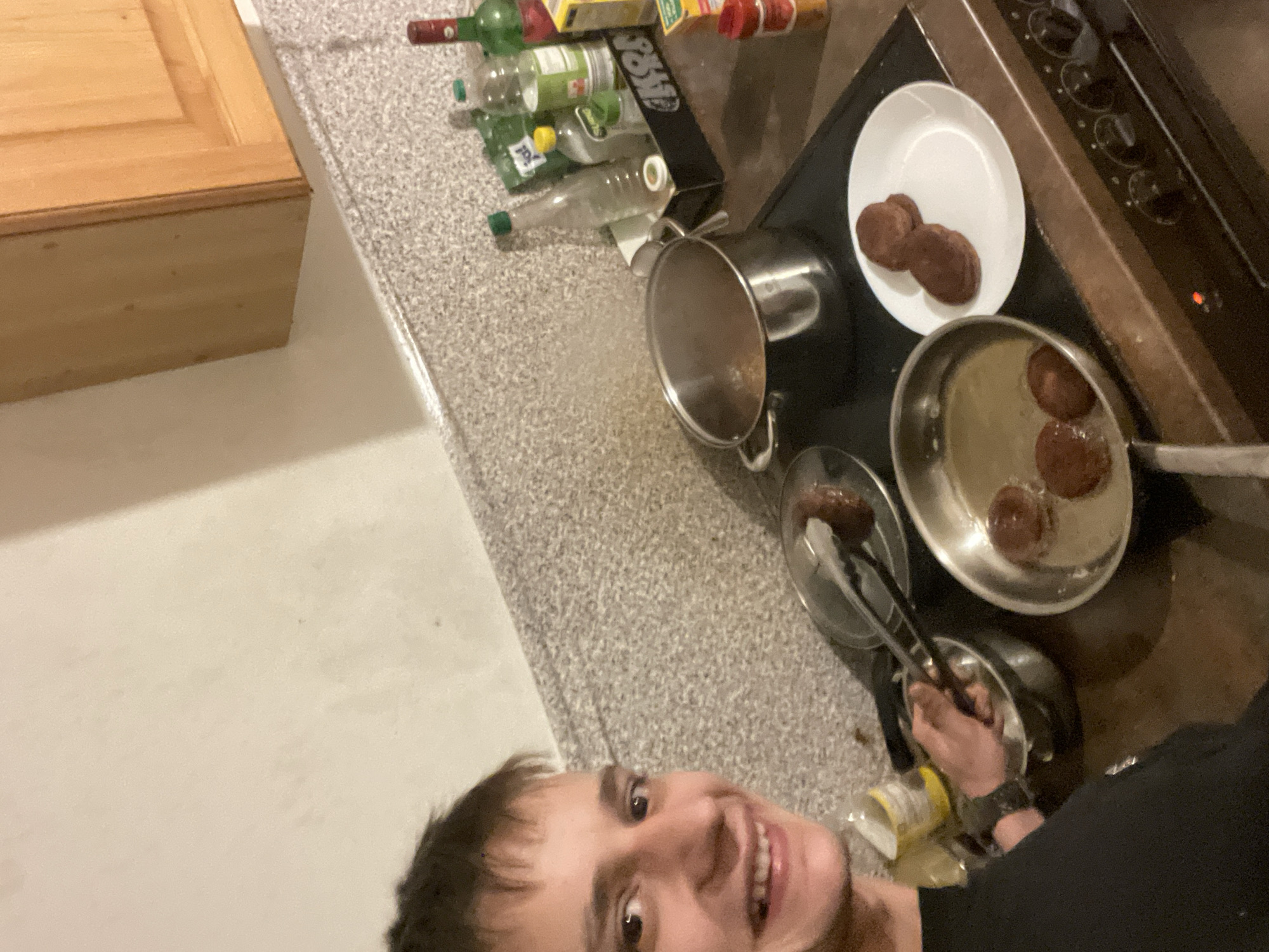 In a kitchen located in Unknown, Unknown, a young male is captured in a close-up selfie while engaged in cooking. The male, appearing to be in his late teens or early twenties, has short, dark brown hair and a fair complexion. He is smiling widely, revealing his teeth, and is wearing a dark-colored short-sleeved top. His left hand is visible, holding a pair of metal tongs over a small silver pot or bowl, suggesting an action related to food preparation. A watch is discernible on his left wrist.
The kitchen environment features a speckled grey and white countertop and light-colored walls. Above the countertop on the right, light brown wooden kitchen cabinets are mounted. A dark-colored electric stovetop with an integrated oven is central to the cooking activity, displaying several control knobs on its front panel and an illuminated red indicator light.
On the stovetop, multiple cooking vessels are present: A large silver pot with its lid partially open is situated on the back-left burner, emitting visible steam. Beside it, the man is manipulating items within a smaller silver pot or bowl. On the front-left burner, a silver frying pan contains liquid, likely oil, and three round, dark brown food items, appearing to be actively frying. To the right, a white ceramic plate holds two similar, already cooked dark brown, round food items stacked one on top of the other. Another silver pan or plate on the back-right burner holds a single dark brown food item, with the tongs resting on it.
Behind the large pot, a black tray organizes various bottles: a clear glass bottle with a green cap, a smaller clear bottle with a yellow cap labeled "SLANG," another clear plastic bottle with a green cap, a green glass bottle with a red cap/seal, and a red-capped bottle with a red label. Additional spice jars or containers, including a red-capped one and a yellow box, are arranged on the right side of the stovetop. A yellow and white container/can is partially visible near the man's left arm.