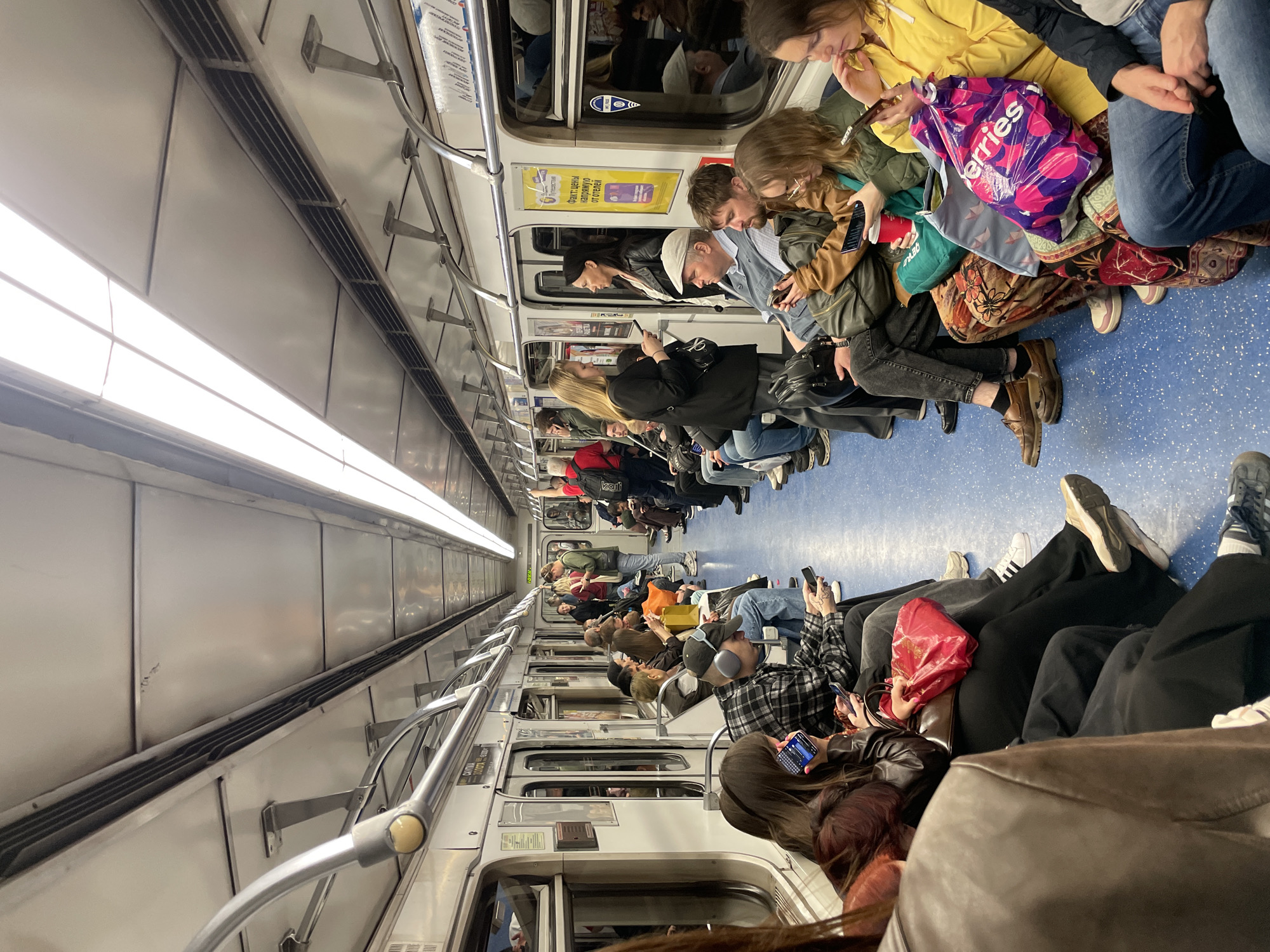 The image shows the interior of a subway car, likely in a major city, as indicated by the densely packed commuters. The subway car is brightly lit by overhead fluorescent lights and features rows of seats on either side of a central aisle. Passengers are seated, most of them preoccupied with their mobile phones. There are various people wearing different styles of clothing. Many people have their legs crossed and are sitting and looking at their phones. The floor is blue. The walls and ceiling of the subway car are white.