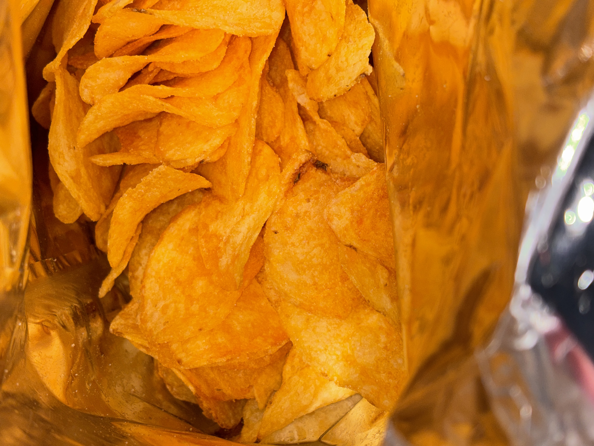 The image shows the interior of an open bag of orange-colored potato chips. The bag is metallic and crinkled. At the top right, the edge of the bag is visible with out-of-focus elements that suggest the bag is resting on a surface, where one can guess as a table. The potato chips are stacked and overlapping, filling the frame. The scene does not give evidence for the location being in the city of Chornomorskoe, Ukraine.