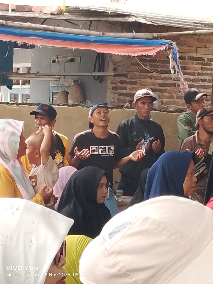 A group of people are gathered outdoors. Several men in the background wear caps, while others wear hijabs in the foreground. One man in a black T-shirt raises his hands, seemingly leading a prayer or discussion. Another man in a dark shirt holds his hands up as if praying, with smoke rising from them. The scene is set against a backdrop of a building, and a colorful banner hangs overhead.