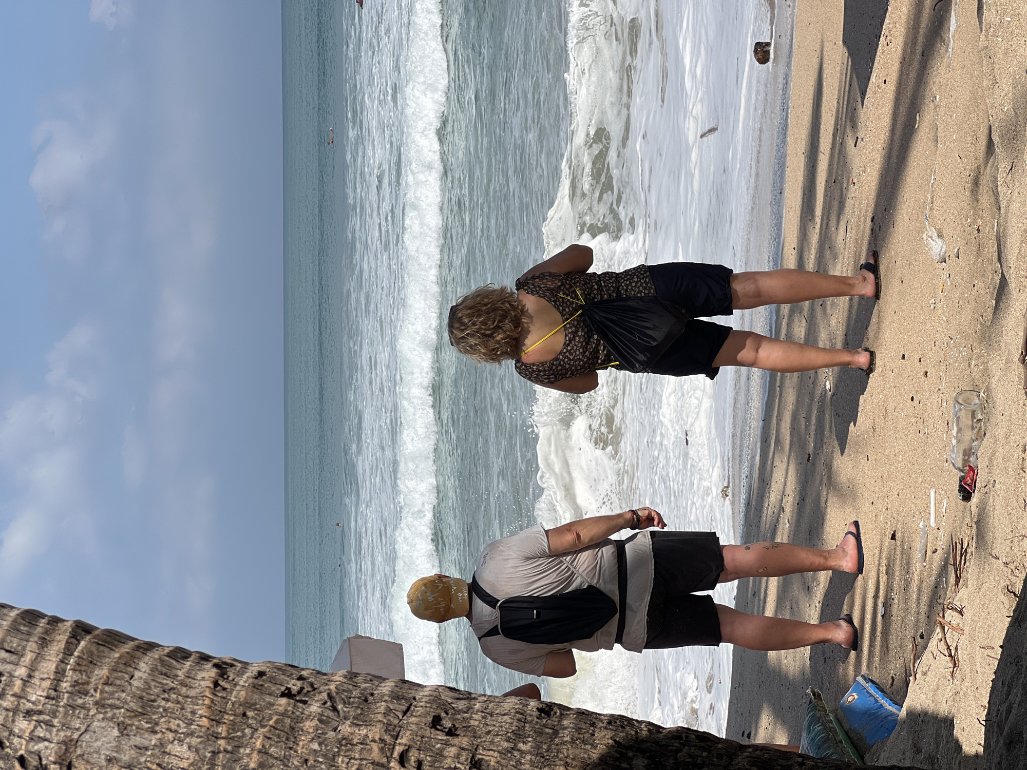 Two individuals stand on a sandy beach, facing the ocean. The person on the left, wearing a light-colored t-shirt, dark shorts, a yellow cap, and a black backpack, is on the left side of the frame. The person on the right, wearing a patterned top, black shorts, and carrying a black bag, is positioned to the right of the first individual. The ocean is visible with waves breaking on the shore. The foreground shows sand with scattered debris. In the background, several people can be seen in the water, with one appearing to be on a surfboard. The sky above is blue with some clouds. The scene appears to be Kuta Beach in Bali, Indonesia.