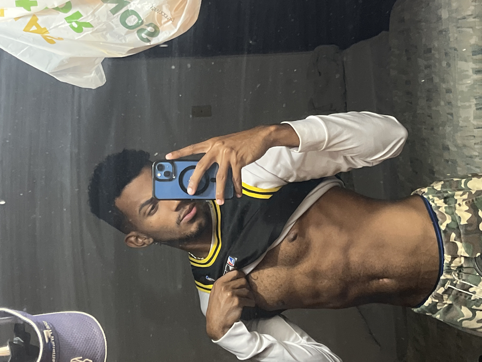 A young man with dark hair is taking a selfie in front of a mirror. He is wearing a black and yellow basketball jersey, a white long-sleeved shirt underneath, and camouflage pants. His torso is exposed. A "Subway" bag is visible in the upper left corner of the frame. A blue and white hat is in the lower left corner. The background appears to be a simple room. The location in the city of Porlamar, Venezuela cannot be determined from the image.