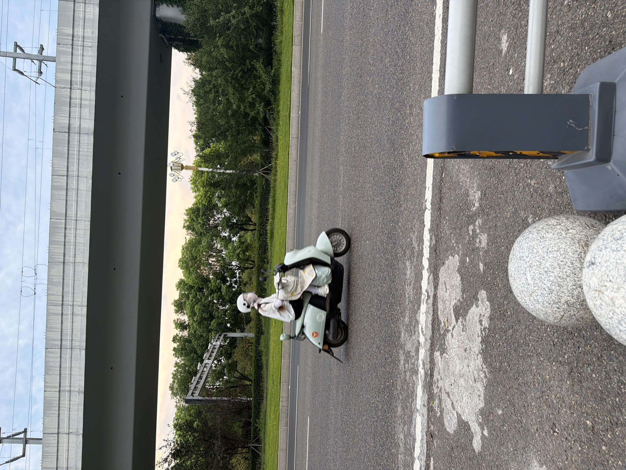 A person wearing a white helmet and face mask rides a light teal scooter down a paved road. The scooter has a white bag hanging from the handlebars and a piece of cloth draped over the front. The person is wearing a white shirt and dark pants. In the background, a concrete overpass runs horizontally across the image, with power lines visible above it. Trees line the road behind the scooter, and a lamppost with multiple lights stands among them. In the foreground, a grey barrier with yellow reflectors and two large, round granite boulders are visible on the side of the road. The scene appears to be set in the city of Jiangjin, China, during the daytime.