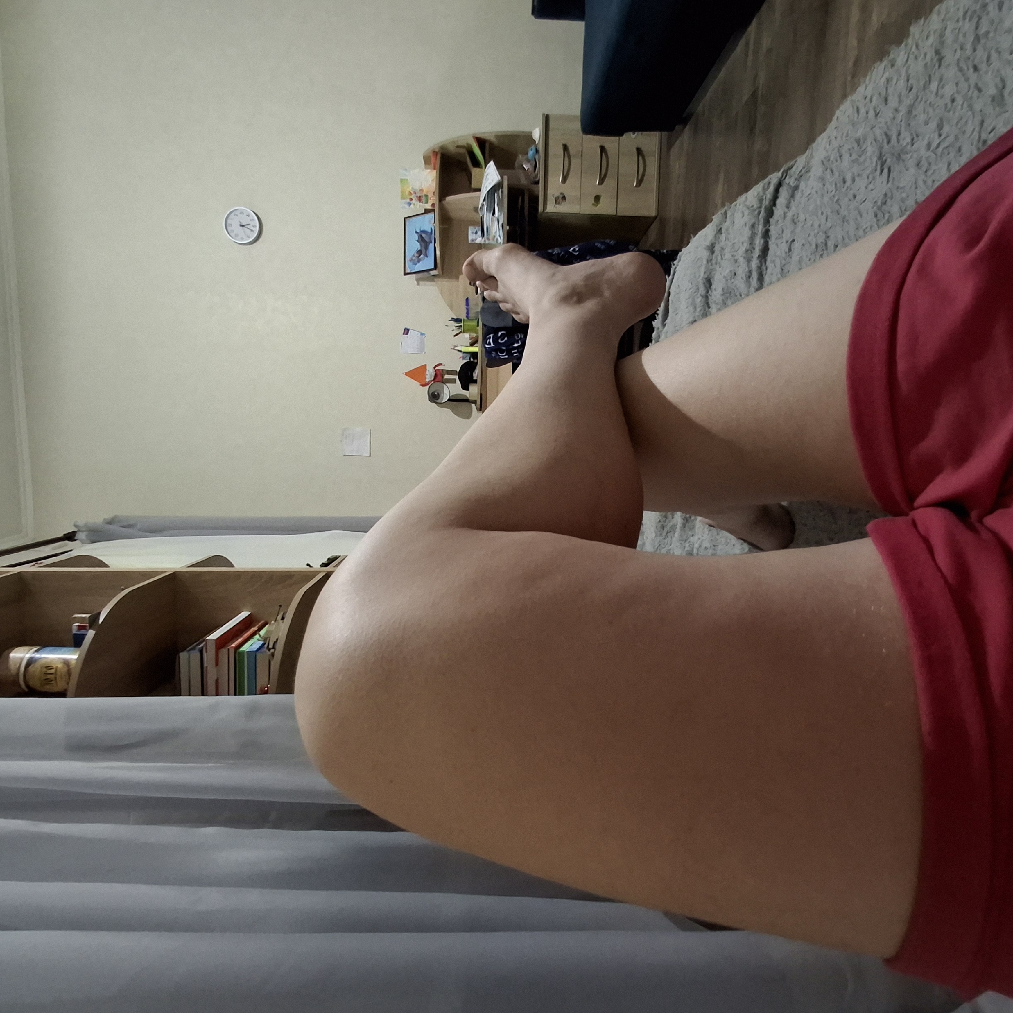 The image shows a person's legs and feet resting on a grey rug. The person is wearing red shorts. In the background, there is a shelf with books and a jar, a desk with a lamp, pens, and papers, and a clock on the wall. There is also a blue couch and a curtain. The scene is indoors, likely in a bedroom or living room. The image is a close-up shot, focusing on the lower body of the person.