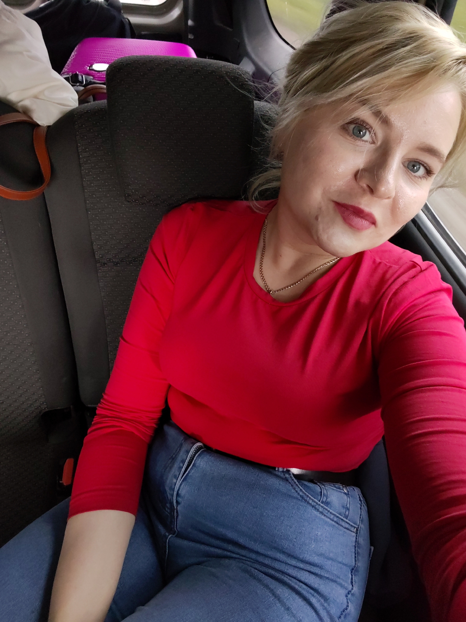 A woman with blonde hair and blue eyes is pictured in the backseat of a vehicle. She is wearing a bright red long-sleeved shirt and blue jeans. Her hair is styled up, and she has on lipstick. She is looking towards the camera, with a slight smile. In the background, a pink suitcase and other luggage are visible, suggesting she is traveling. The interior of the vehicle, including the seat upholstery, is dark in color. A hint of the outdoors, possibly a road, is visible through the window on the right side of the frame. The lighting suggests it is daytime. The image appears to be a selfie taken by the woman. Based on the provided context, the location is Buranovo, Russia.