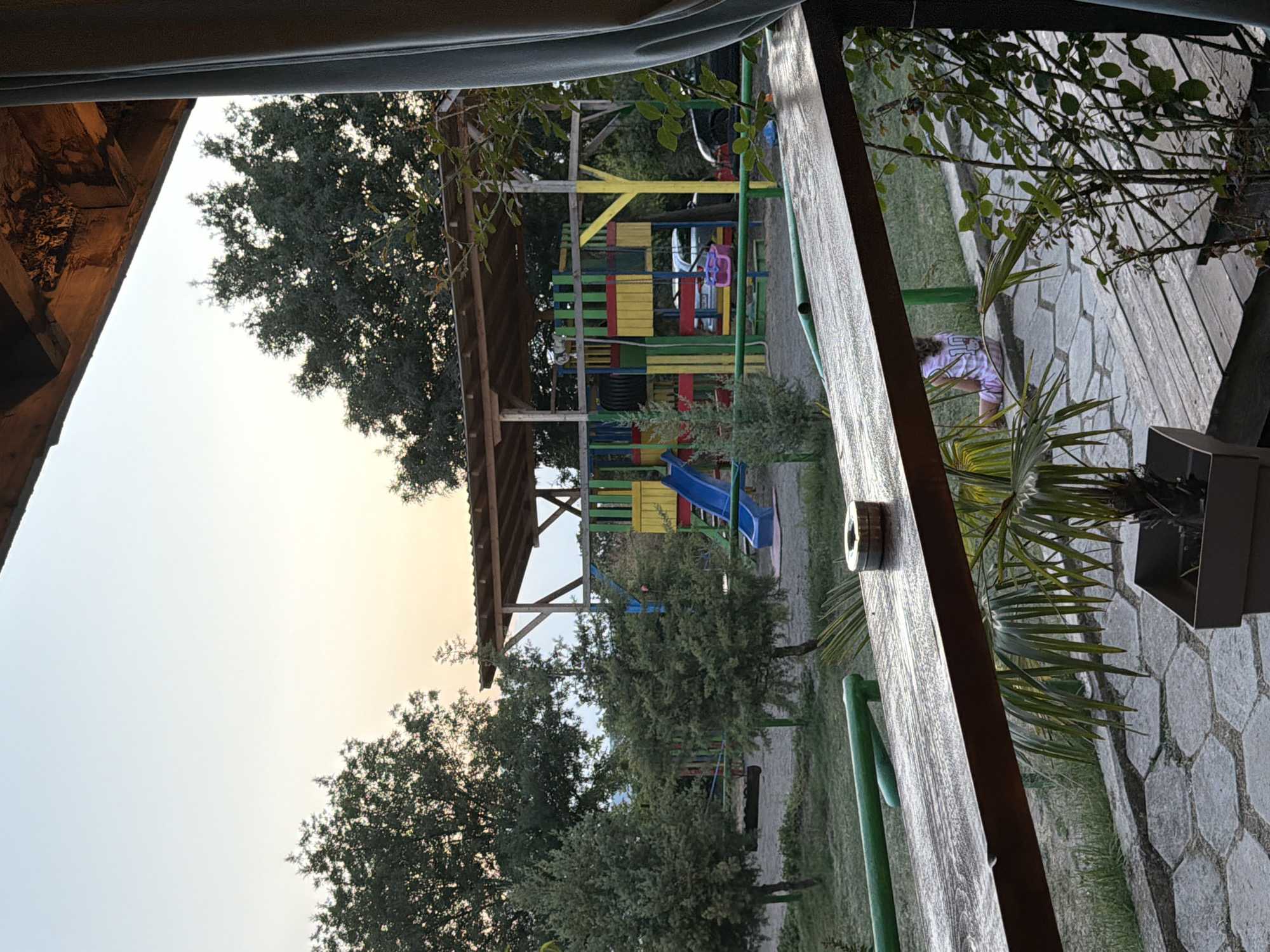The image depicts an outdoor scene, likely a backyard or park area, with a playground structure in the background. In the foreground, a wooden surface, possibly a table or railing, runs across the frame, with a metal ashtray placed on its left side. To the right of the wooden surface, a small potted plant with palm-like fronds is visible. A person, appearing to be a child, is seated on the ground to the right, facing away from the camera, wearing a light-colored garment with a design on the back. The playground features colorful slides, swings, and climbing structures, all under a wooden-roofed canopy. Trees and greenery surround the playground. The sky in the background suggests either dawn or dusk. There are no identifiable landmarks of Tvarditsa, Bulgaria in the image.