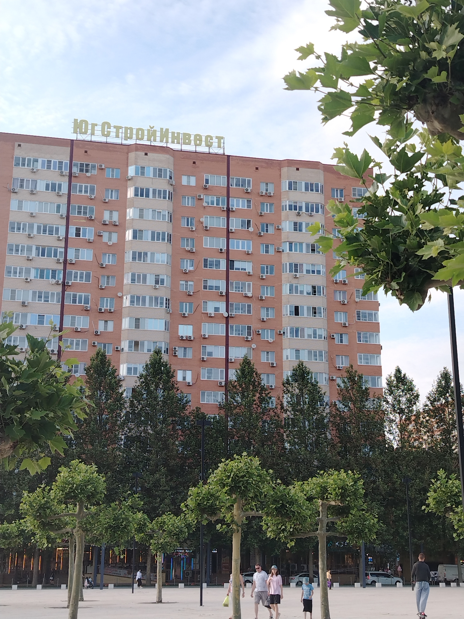 The image shows a large, multi-story brick apartment building with many windows and air conditioning units. A sign with yellow lettering spelling "ЮгСтройИнвест" is visible at the top of the building. In the foreground, there are several manicured trees and a paved area where a few people are walking and riding a scooter. Some cars are parked in the background. The sky is cloudy. This image was taken in Krasnodar, Russia.
