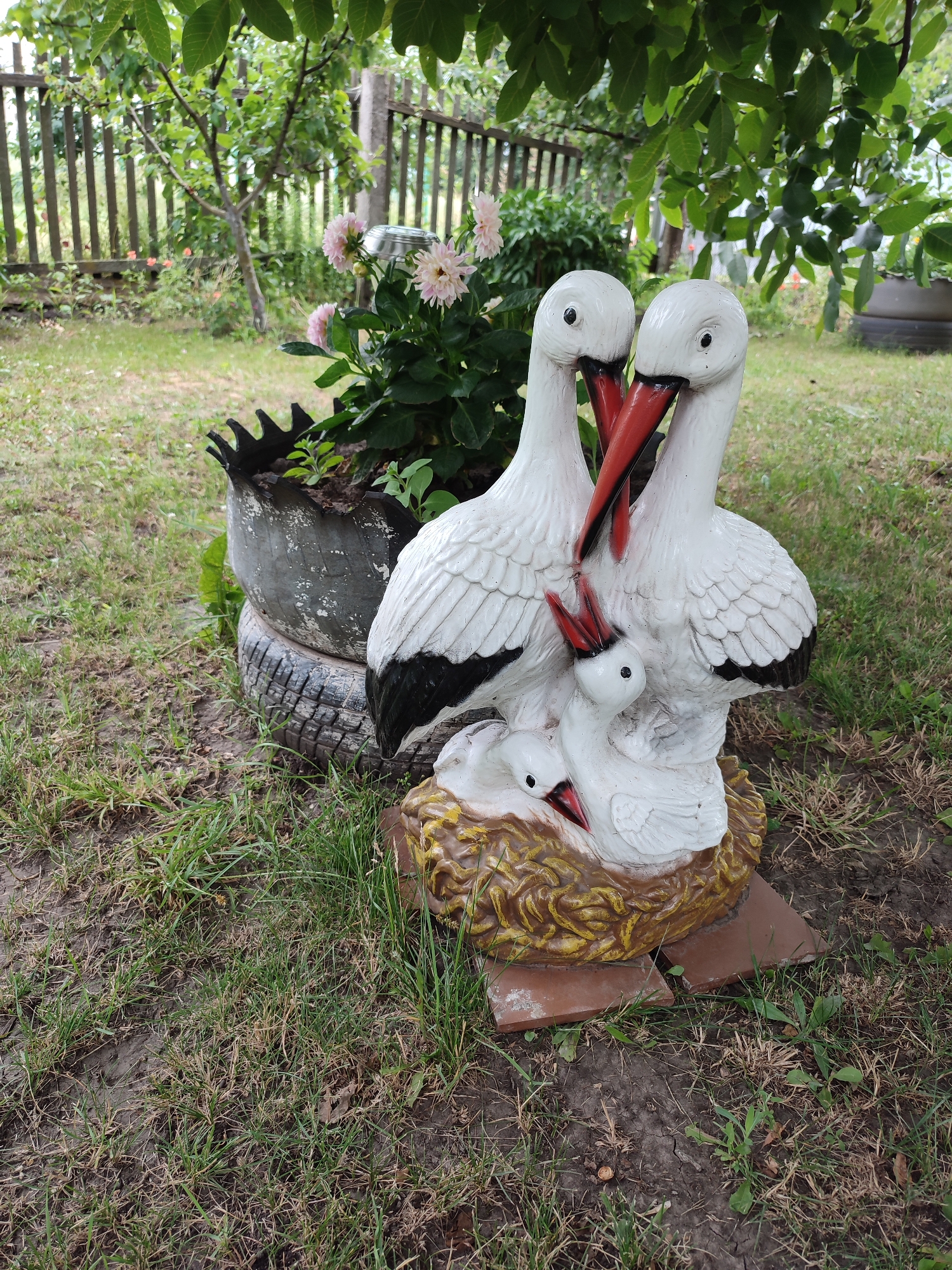 In Voskobiinyky, Ukraine, a garden scene features a white ceramic sculpture of storks family placed in a nest. The sculpture depicts two adult storks and two chicks, nestled together. The adult storks have red beaks and black wing tips. Behind the sculpture, a wooden fence encloses a garden with green grass and a bush with pink flowers. The sculpture is positioned on a base of broken terracotta tiles. The surrounding environment includes dirt paths and dry grass, suggesting a natural, outdoor setting.