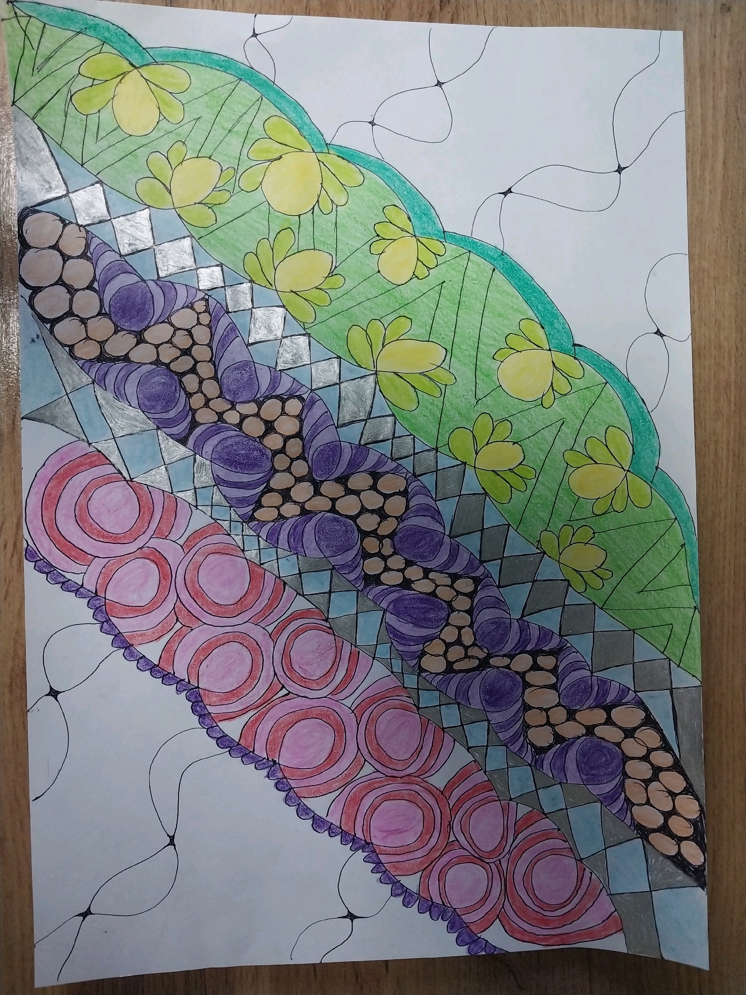 This is a close-up, overhead shot of a hand-drawn Zentangle-inspired artwork on white paper. The artwork is colored with colored pencils and features a repeating pattern of curved bands filled with different designs. From top to bottom, the bands are:
1. A curved green band with light yellow flower-like shapes.
2. A band with a checkerboard pattern of light blue and gray squares, interspersed with a section of circular, brown shapes.
3. A band filled with wavy purple lines and brown circular shapes.
4. A band with pink and red concentric circles.
5. A scalloped purple border along the bottom edge of the artwork.
The entire artwork is framed by a loosely drawn net-like pattern of black lines. The paper is resting on a wooden surface, likely a table. The lighting suggests an indoor setting. The image does not depict any specific location in Dnipro, Ukraine.