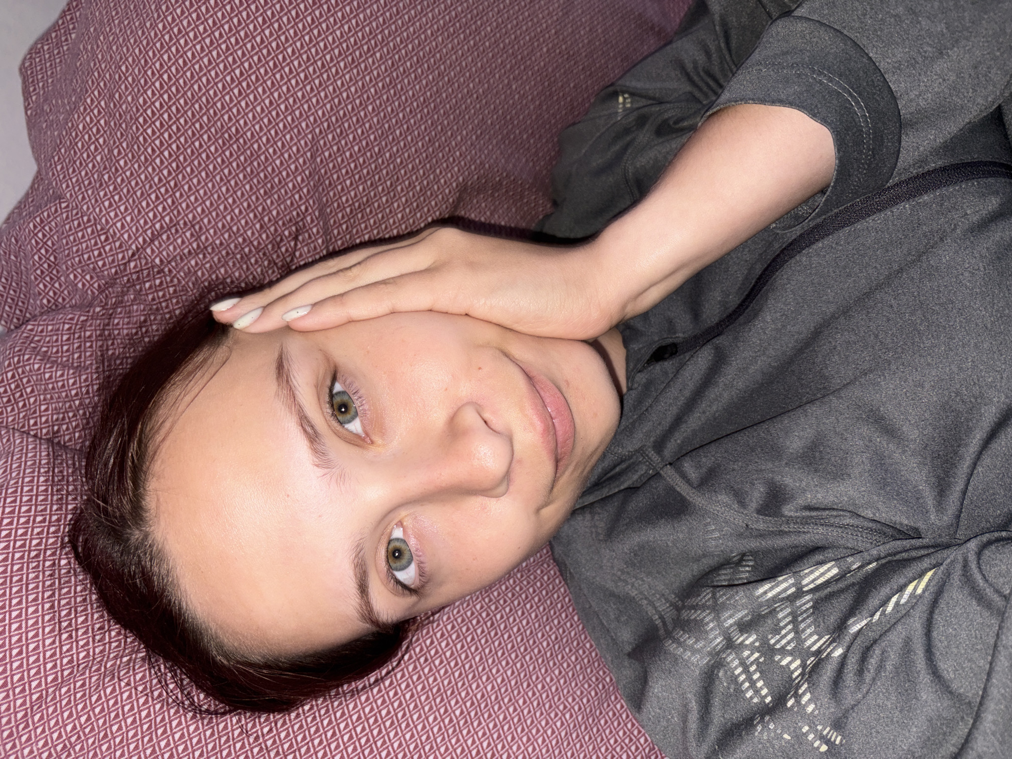 A young adult female with dark hair and green/hazel eyes is depicted in a close-up, top-down shot. She is lying down, possibly on a bed, with her head resting on a reddish-maroon colored fabric featuring a white geometric diamond pattern. She wears a dark grey athletic jacket with a zippered front and a subtle light-colored pattern on the left shoulder/sleeve. Her right hand is placed against her right cheek, with fingers slightly spread. Her fingernails are painted white with small dark specks. She exhibits a slight smile and looks directly at the camera. The exact location in the city of Прикамский, Russia, cannot be determined from the visual information present in the image.