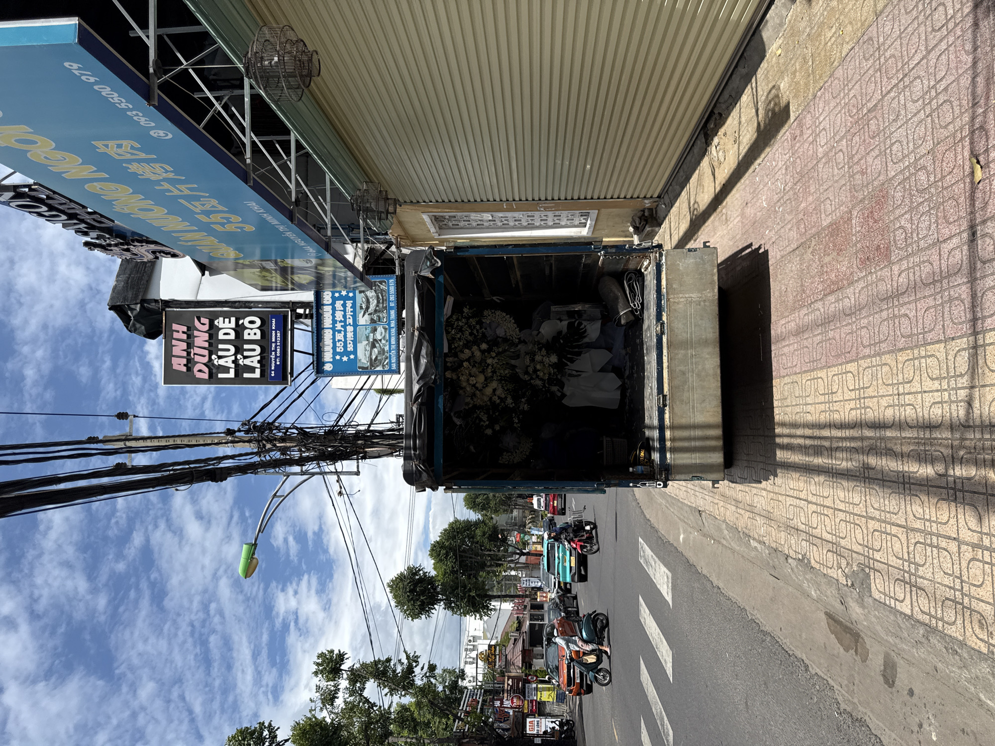 The image depicts a street scene in Nha Trang, Vietnam. In the foreground, a blue truck is parked, its rear open to reveal a large arrangement of white flowers, likely for a funeral. A sidewalk with patterned tiles runs alongside the street. On the street, several vehicles are present, including scooters and cars, indicating moderate traffic. Pedestrians are visible on the scooters, one of whom is wearing a light-colored shirt and pants, and another in a dark blue shirt and white pants. Overhead, numerous electrical wires crisscross the sky, along with streetlights. Signs with Vietnamese text are visible on buildings lining the street. The sky is partly cloudy. The overall impression is a busy urban environment with a somber element in the truck's cargo.
