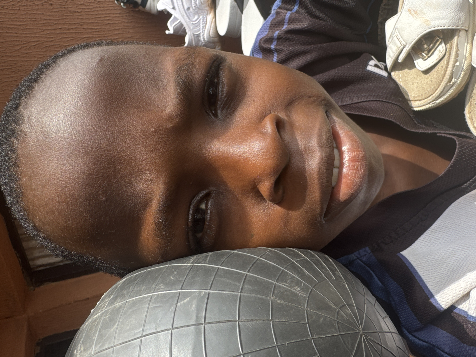 A close-up, tilted shot depicts a Black child resting their head on a soccer ball. The child has short, dark hair and dark eyes. Their lips are slightly parted, revealing a hint of teeth. They are wearing a dark shirt with blue and white stripes. In the blurred background, there are white sneakers and sandals. The location is specified as Onitsha, Nigeria, but there is no visual evidence in the image to confirm this.