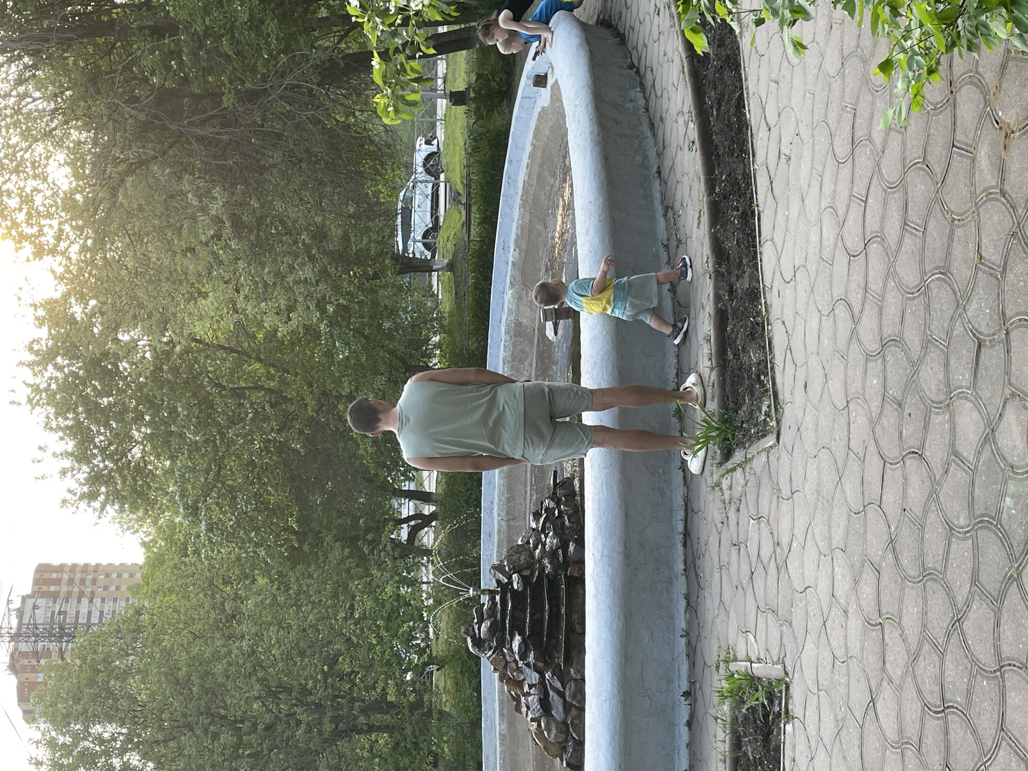 The scene captures an outdoor setting, likely within a park in Perm, Russia, considering the city's skyline visible in the background. A concrete fountain forms the central element, with tiered rock formations from which water spouts. A man wearing a light green tank top and shorts stands facing the fountain, seemingly observing the water feature. To his right, a young child in a yellow and blue shirt and gray shorts is also positioned near the fountain, possibly interacting with the water. Further along the fountain's perimeter, two more children are present, peering into the water. Paved walkways surround the fountain, exhibiting a geometric pattern of interlocking shapes. The environment is lush with mature trees providing shade and contributing to a natural atmosphere. Several cars are parked on the street in the background.