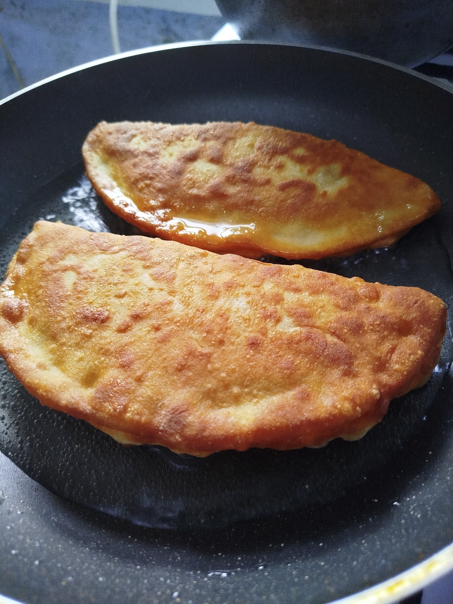 Two golden-brown, pan-fried pasties are cooking in a black frying pan filled with oil. The pasties are crescent-shaped and appear to be filled with a light-colored ingredient. The pan is on a stovetop, and the surrounding environment is out of focus but suggests a kitchen setting. The image was taken in Ceadîr-Lunga, Moldova.