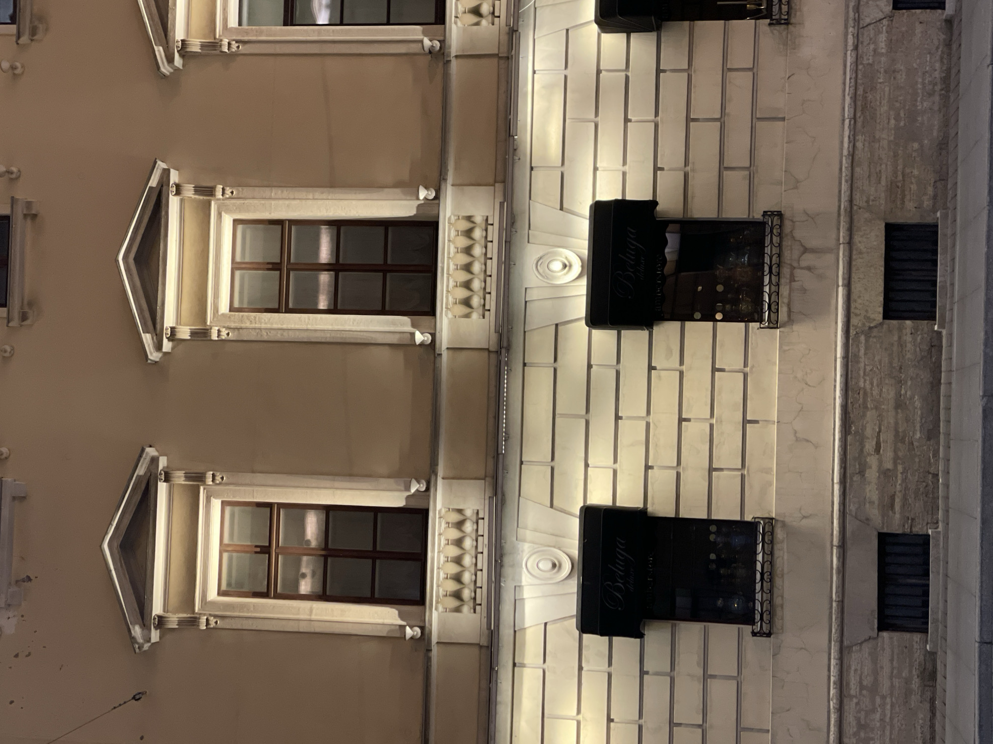 The image captures a section of a building facade at night, likely in Saint Petersburg, Russia, based on the architectural style. The building has a mix of beige and white stone cladding. There are two prominent windows with decorative triangular tops on the left side of the image. Adjacent to these windows, a row of teardrop-shaped decorations separates the beige section from a white section. On the right, two dark awnings with "Beluga" written on them are visible. The white section of the facade is patterned with rectangular blocks. Further to the right, another section of the building has a rough, textured stone surface with smaller windows. The image is taken at night, and the building is illuminated by artificial lighting. There are no people visible in the shot.