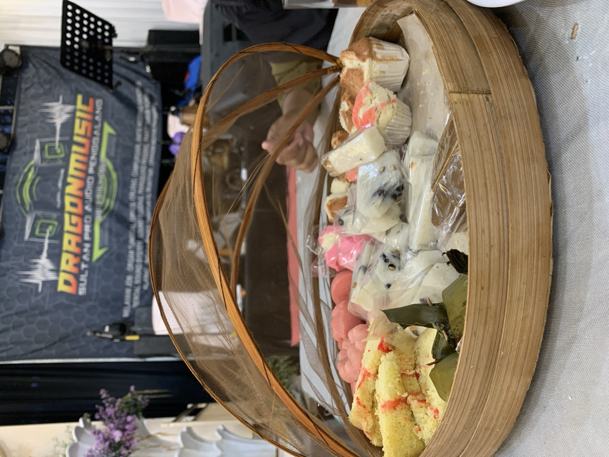 The image shows a basket of assorted pastries, likely local Indonesian snacks, covered with a mesh food cover. The basket is placed on a table with a light-colored tablecloth. In the blurred background, there is a banner for "DRAGON MUSIC PRO AUDIO PENGGALANG SULTAN PR", indicating a music-related event or establishment in Desa Penggalang Kecamatan Adipala, Indonesia. Floral decorations are visible in the foreground. The pastries include cupcakes, sliced cakes with red accents, and other individually wrapped sweets. The lighting appears to be indoor, likely during an event or gathering.