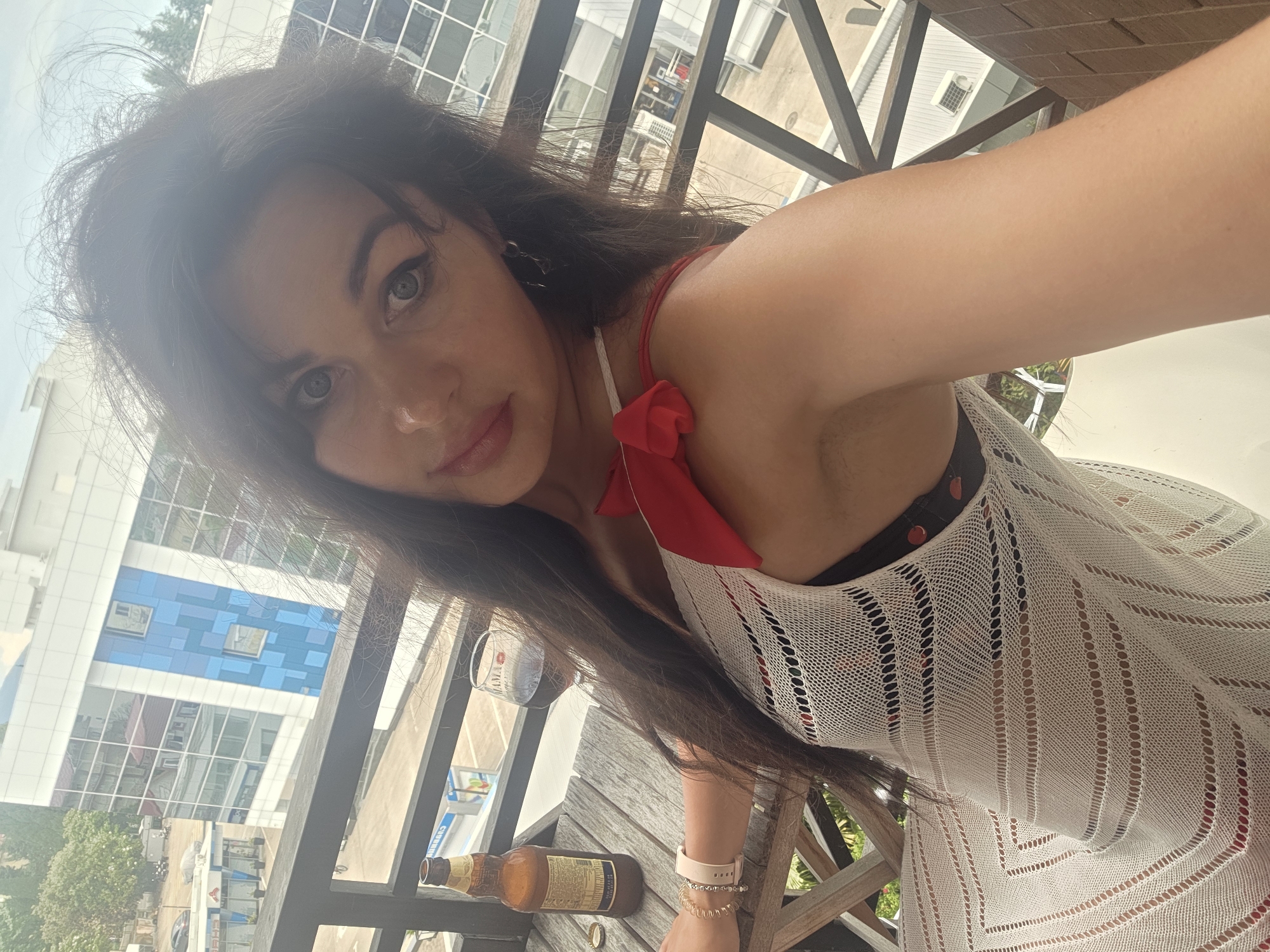 A woman is taking a selfie on a balcony overlooking a city street in Sochi, Russia. She has long dark hair and is wearing a white open-knit dress over a black and red patterned bikini. There is a bottle of beer and a glass of red wine on a wooden table next to her. The background shows a modern building with blue and white panels, trees, and a cloudy sky.
