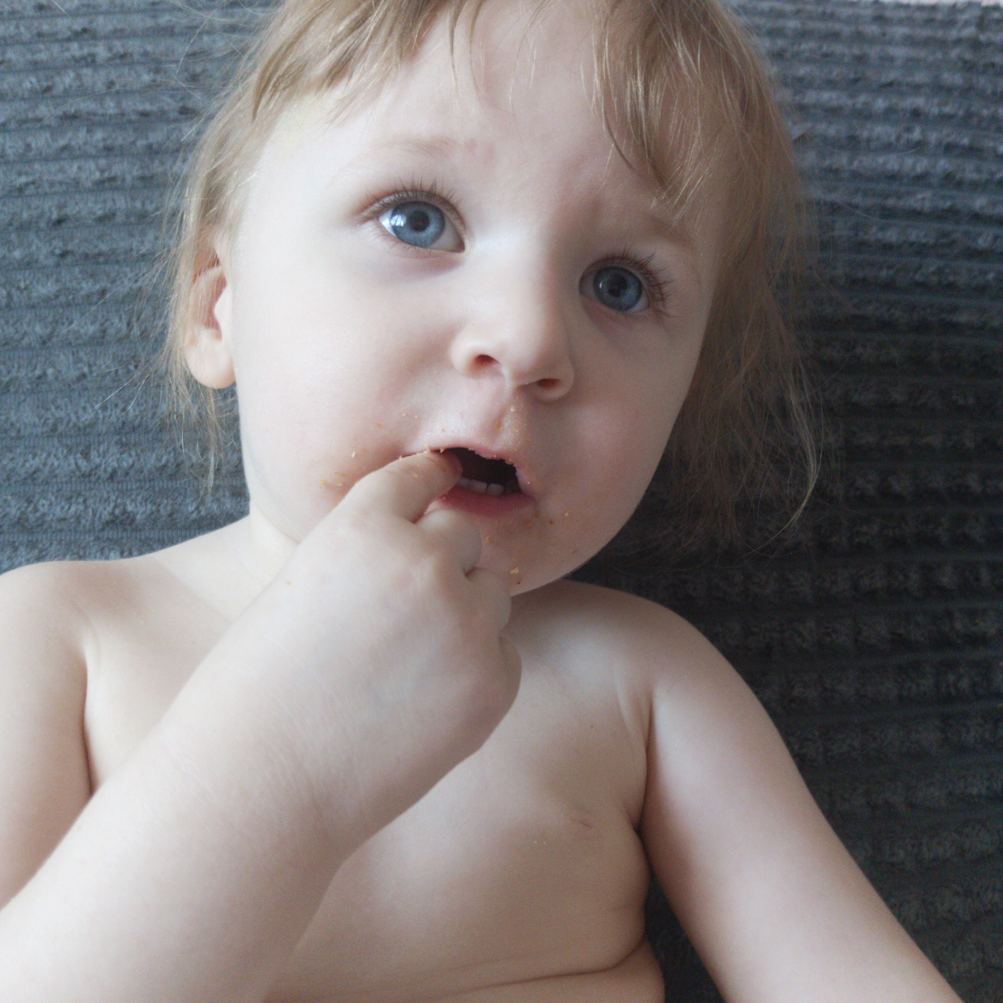 A close-up, eye-level shot in the city of Chaikovsky, Russia, shows a fair-skinned toddler with light blonde hair and blue eyes, positioned against a dark, horizontally ridged textured background. The child is shirtless, with their right index finger partially inserted into their open mouth. Visible around the child's mouth, on the chin, lower lip, and philtrum, are scattered yellowish-brown particulate matter, consistent with food crumbs. The child's gaze is directed slightly upwards and to the right of the frame, with an open-mouthed expression.