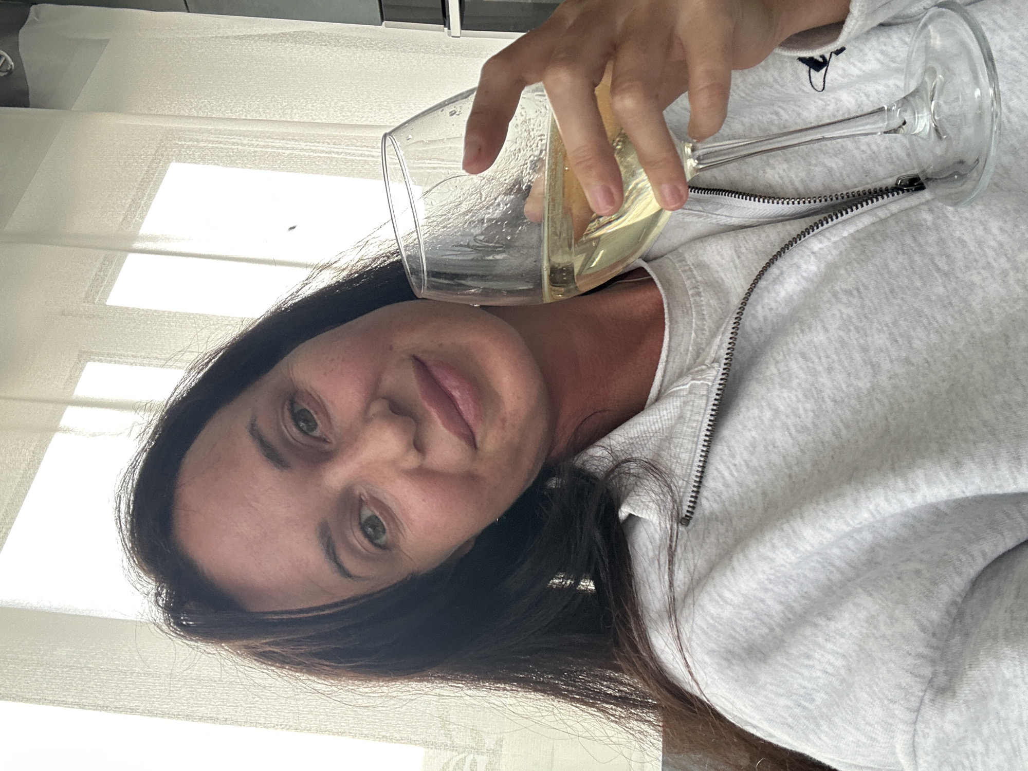 A woman with long dark hair and fair skin holds a stemmed glass of white wine. She is wearing a light grey zippered jacket over a white t-shirt. The background shows bright windows with sheer white curtains. The image appears to be a selfie taken indoors. The provided location of Baklashi, Russia, is not discernible from the visual information in the image.