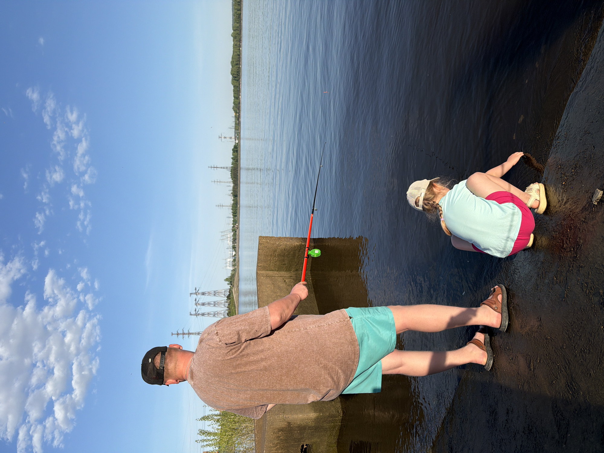 A man and a girl are fishing from a concrete structure near a body of water. The man, wearing a brown t-shirt, green shorts, and sandals, holds a fishing rod with a red handle and a float. The girl, wearing a light blue shirt, pink shorts, and a hat, is crouched down near the water's edge. The water is calm, and the sky is blue with some clouds. Transmission towers are visible in the background. The scene appears to be in Chaikovsky, Russia, based on the possible scenery.