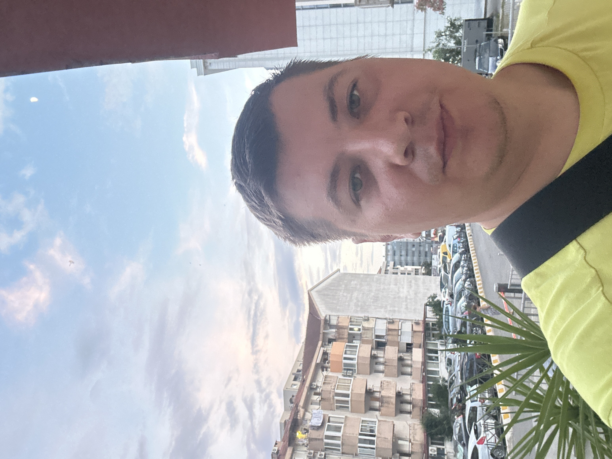 This is a selfie taken by a person wearing a yellow shirt, with a black strap across their shoulder. The person is looking towards the camera with a neutral expression. The background shows a city scene with buildings and parked cars. The sky is visible, with clouds suggesting it might be late afternoon or early evening. The image is oriented sideways, so the man's head is on the right side of the frame. The setting appears to be an urban environment, possibly a street or parking lot in Budva, Montenegro, given the building architecture. There is no visible text in the image.