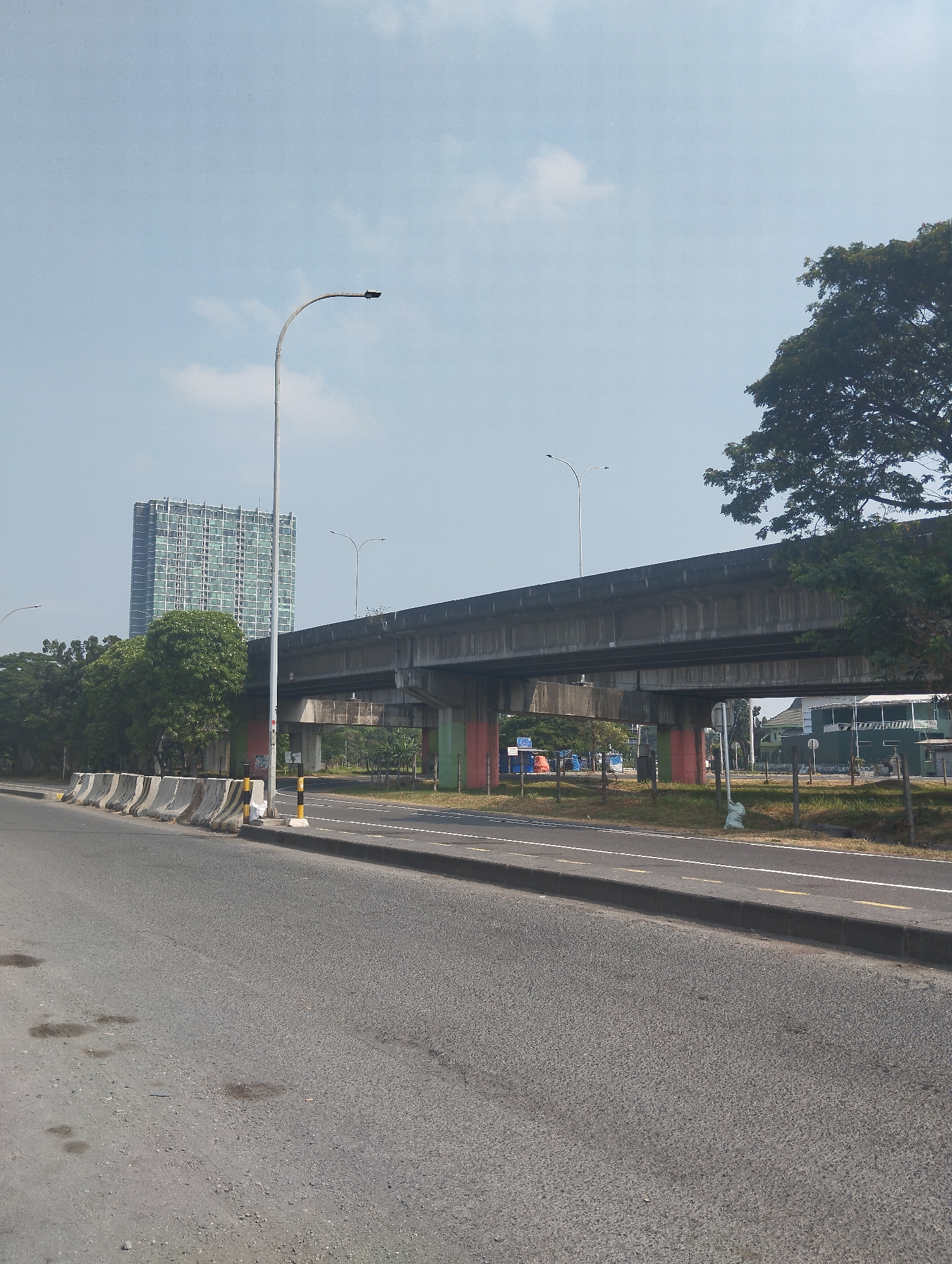 A wide, asphalt road runs diagonally across the foreground of the image, from the lower left to the mid-right. The road appears to be an urban highway or major thoroughfare, with distinct lanes marked by white and yellow lines. The road surface shows signs of wear, with several dark patches that suggest water stains or oil spills. Along the left side of the road, a series of concrete barriers are placed at intervals, indicating a separation from an adjacent area.
Dominating the mid-ground is a raised concrete overpass, its underside supported by sturdy concrete pillars. These pillars are painted with vertical bands of red and green. The overpass itself is a broad, flat structure, suggesting it carries a significant amount of traffic, though no vehicles are currently visible on it.
Beyond the overpass, a tall, modern building with a grid-like facade of windows rises into the sky. This building appears to be a skyscraper or a large office complex. To the right of the building, lush green trees form a dense canopy, partially obscuring the view of the sky. A large, mature tree with dark green foliage is prominent in the upper right corner of the image, its branches extending over the scene.
The sky is a pale, hazy blue with a few wispy white clouds. The overall lighting suggests it is daytime, possibly late morning or early afternoon, with a moderate level of brightness. The environment appears to be a mix of urban infrastructure and natural elements, characteristic of a developing city.
The scene can be identified as being located in Surabaya, Indonesia, specifically along the road leading to or from the Juanda International Airport. The presence of the modern high-rise building, the elevated road infrastructure, and the characteristic tropical vegetation are all consistent with this location.