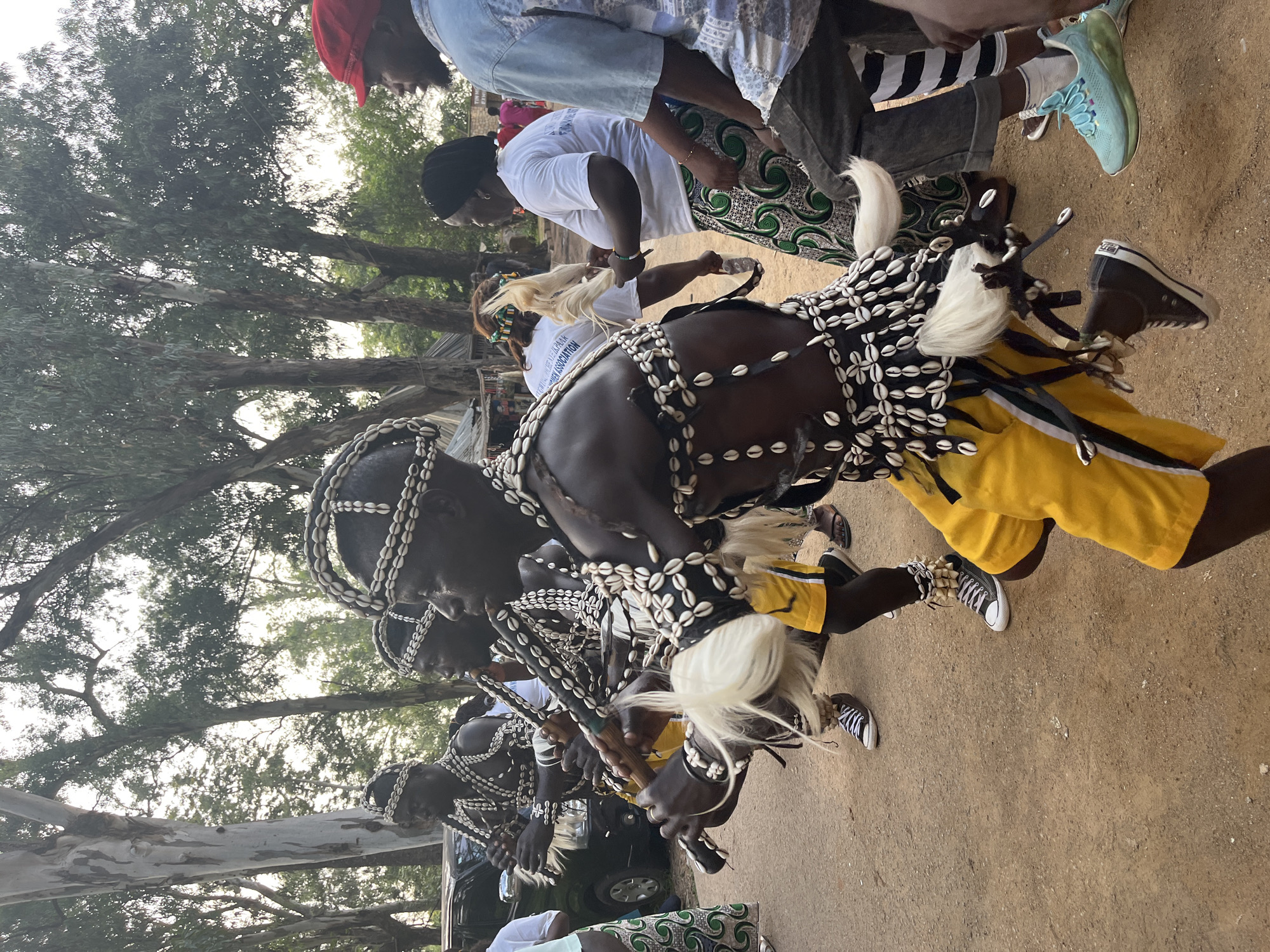 The image captures a cultural performance in the city of Jos, Nigeria, possibly during a festival or celebration. The foreground is dominated by several men in traditional attire. They are adorned with elaborate necklaces, bracelets, and headdresses made of shells and beads. Their bodies are painted, and they wear brightly colored yellow shorts and black leggings. Two of the men are playing flutes, their movements suggesting a rhythmic dance. One of them has white fur or hair attached to his costume. Behind them, several onlookers are visible, some in casual clothing and others in traditional wear. The background features tall trees, characteristic of the Jos Plateau's landscape, and a glimpse of a vehicle, suggesting an outdoor event. The ground is dusty and appears to be a dirt path or open space. The lighting indicates it is likely daytime, possibly late afternoon given the soft light.