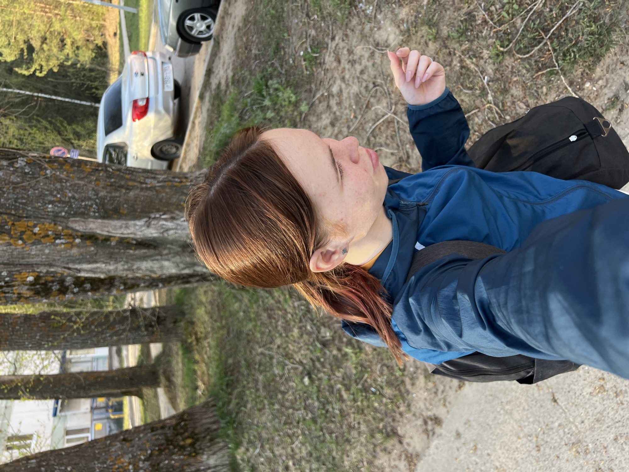 A medium shot depicts a woman outdoors, likely in Chaikovsky, Russia. She stands on a patch of ground interspersed with dirt and sparse greenery, adjacent to a paved area that may be a road or sidewalk. Parallel to the woman and ground are a line of mature trees with textured bark, which create a blurred backdrop obscuring buildings in the distance. Partially visible between the trees is a light-colored car with a discernible license plate. The woman wears a blue jacket and has a black backpack slung over her shoulder. Her hair is brown with some reddish highlights. Her right hand is raised, and her face is in profile, facing slightly upwards.