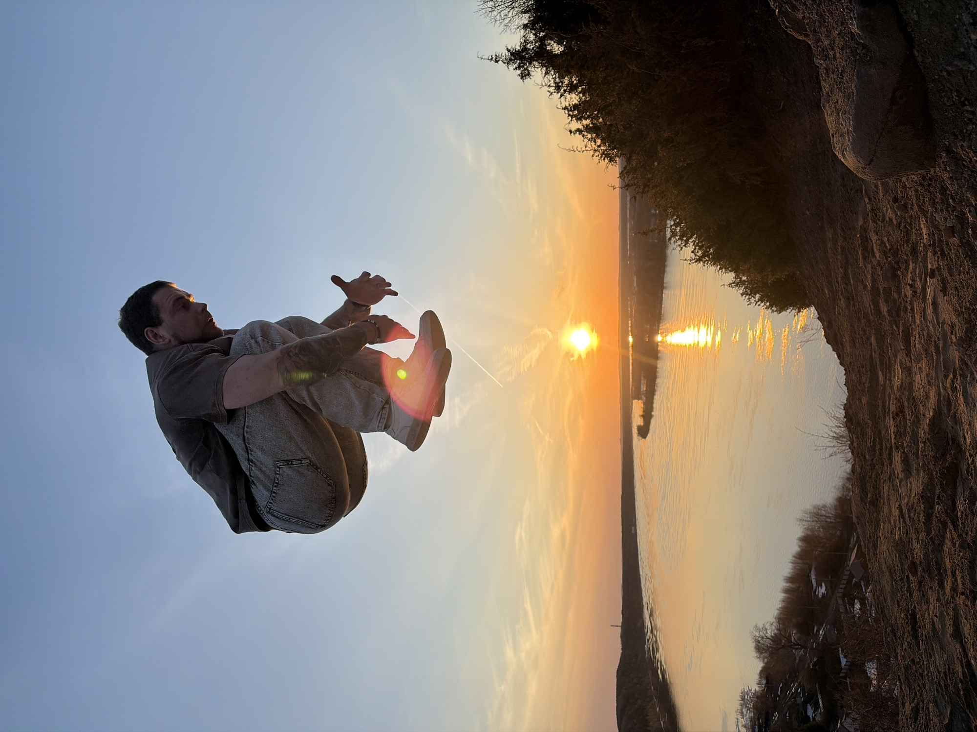 The image depicts a man in mid-air, seemingly performing a backflip, with a sunset over water in the background. He is wearing a short-sleeved shirt and jeans, with his legs tucked up towards his chest and arms extended. The sunset reflects on the water, creating a bright, golden glow. Landmass with trees is visible to the right of the frame. The location appears to be Ольховка, Russia, based on the surrounding environment and the sunset over water.