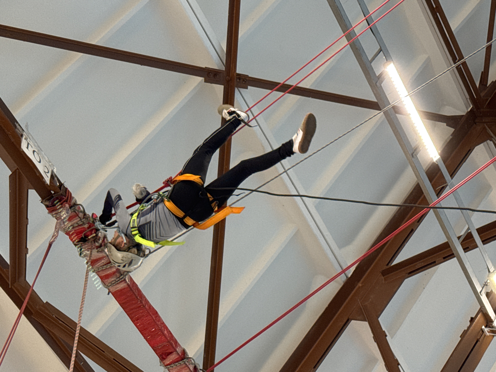 A person wearing a helmet and safety harness is suspended upside down from a red-wrapped structural beam via ropes and cables within an indoor space. The individual is wearing black pants, a gray shirt with black accents, and white sneakers. The ceiling has a repeating triangular pattern with brown metal supports. A bright white light fixture is visible. The location is in the city of Unknown, Unknown.