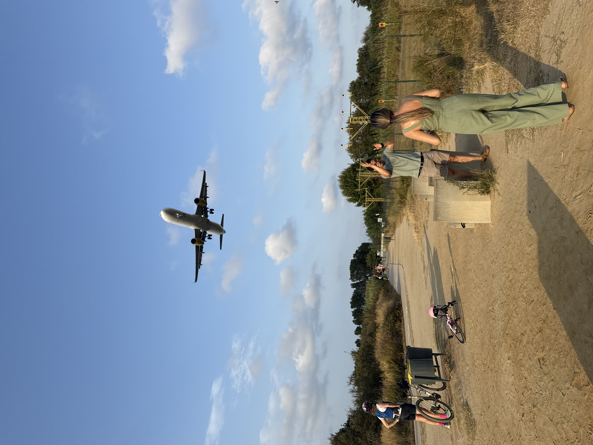 An airplane is flying in a clear blue sky with scattered clouds over a sandy path in El Prat de Llobregat, Spain. On the left of the path, a woman in a blue and white athletic top and black shorts stands with her hand on her hip, next to a bicycle and a trash bin. A child's bicycle with a pink helmet lies on the ground near the woman. Further down the path, a man in a green t-shirt and brown shorts is gesturing with his hands, and a woman in a green jumpsuit is facing away from the camera, also looking up at the sky. In the background, there are trees, dry grass, and airport landing lights.