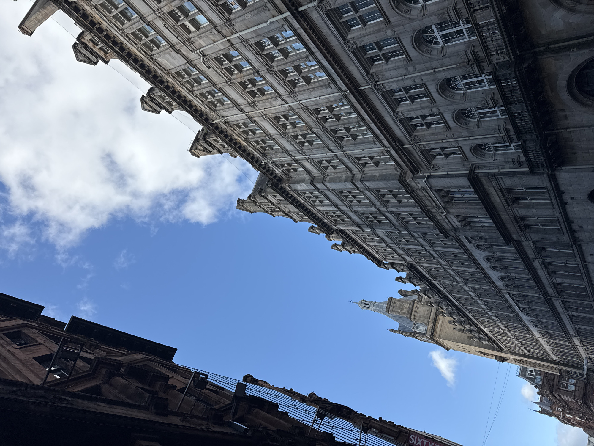 This is a vertical, wide-angle, low-angle shot looking up at the sky between two buildings in Glasgow, Scotland. The sky is a vibrant blue with scattered white clouds.
The building on the left is made of dark reddish-brown stone with ornate architectural details, including what appear to be decorative cornices and window frames. The building on the right is much larger and constructed of grey stone. It features a repeating pattern of numerous windows, many with arched tops, and elaborate decorative elements. Part of a clock tower or spire is visible in the distance, also made of stone and topped with a pointed roof.
The perspective emphasizes the grandeur and height of the buildings, creating a sense of looking up from a narrow street. The angle also suggests that the photograph was taken on a clear day, likely in the daytime, given the bright sky and lighting on the buildings. There is a sign visible at the bottom left corner with the text "SIXTY". The scene evokes a sense of urban grandeur and architectural history.