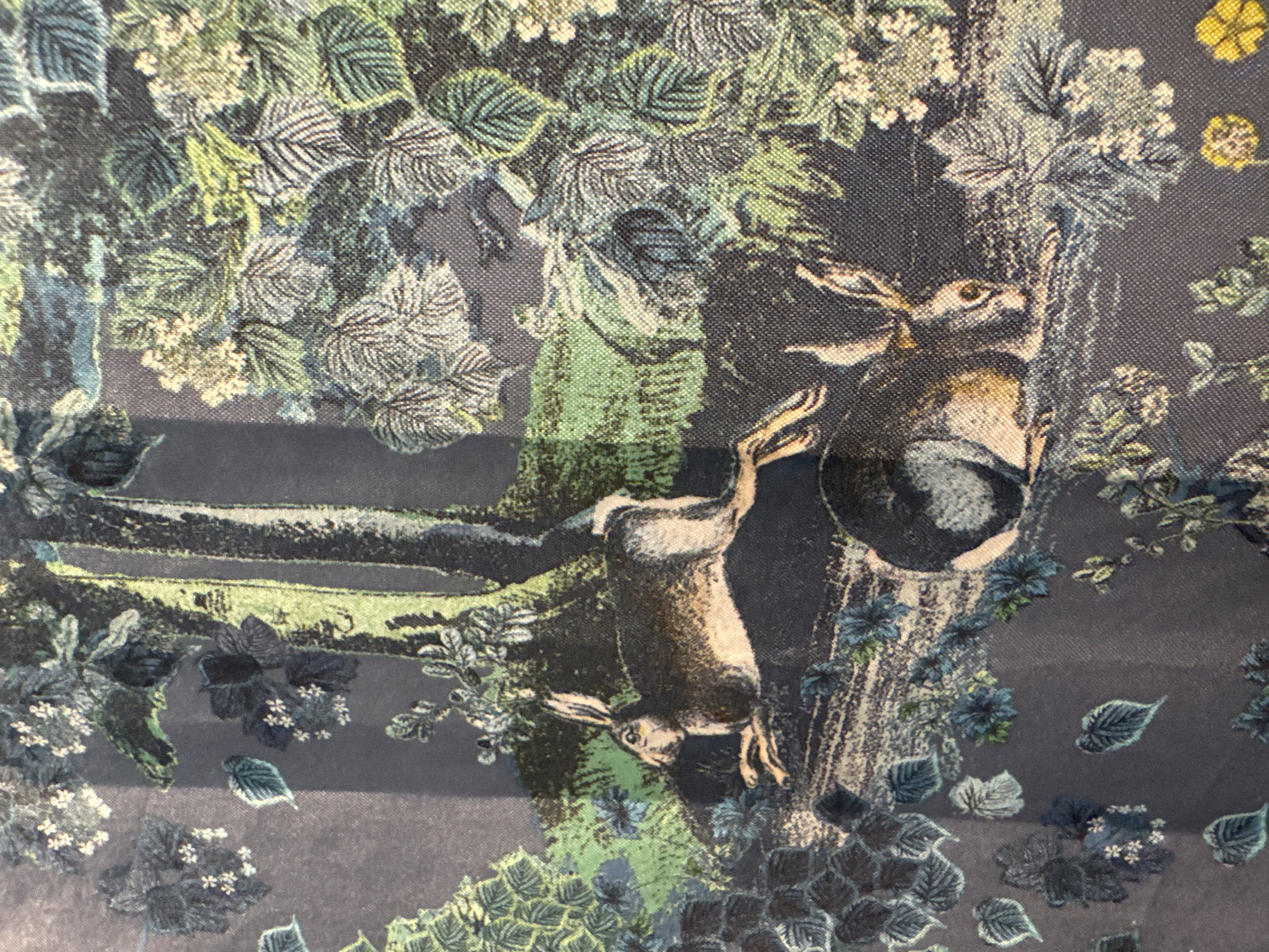 The image is a close-up, low-angle shot of a fabric print depicting a woodland scene with two hares. The fabric has a dark, grayish-blue base color. The print features a stylized forest environment with trees, foliage, and small white flowers.
In the foreground, slightly to the left, a hare is depicted in a crouched position, facing left with its head slightly lowered. It has brown fur with lighter underparts and visible hind legs. Behind and slightly to the right of the first hare, another hare is shown lying down, facing towards the right. This hare also has brown and white fur, with its hind legs tucked underneath.
The background is filled with dense foliage rendered in shades of green, blue, and white, with hints of light brown in the tree trunks. The style of the print is somewhat illustrative, with fine details in the rendering of the leaves and the hares. The texture of the fabric is visible throughout the image, suggesting it might be a woven material like linen or cotton.
There is no indication of a specific city or location within the image itself.