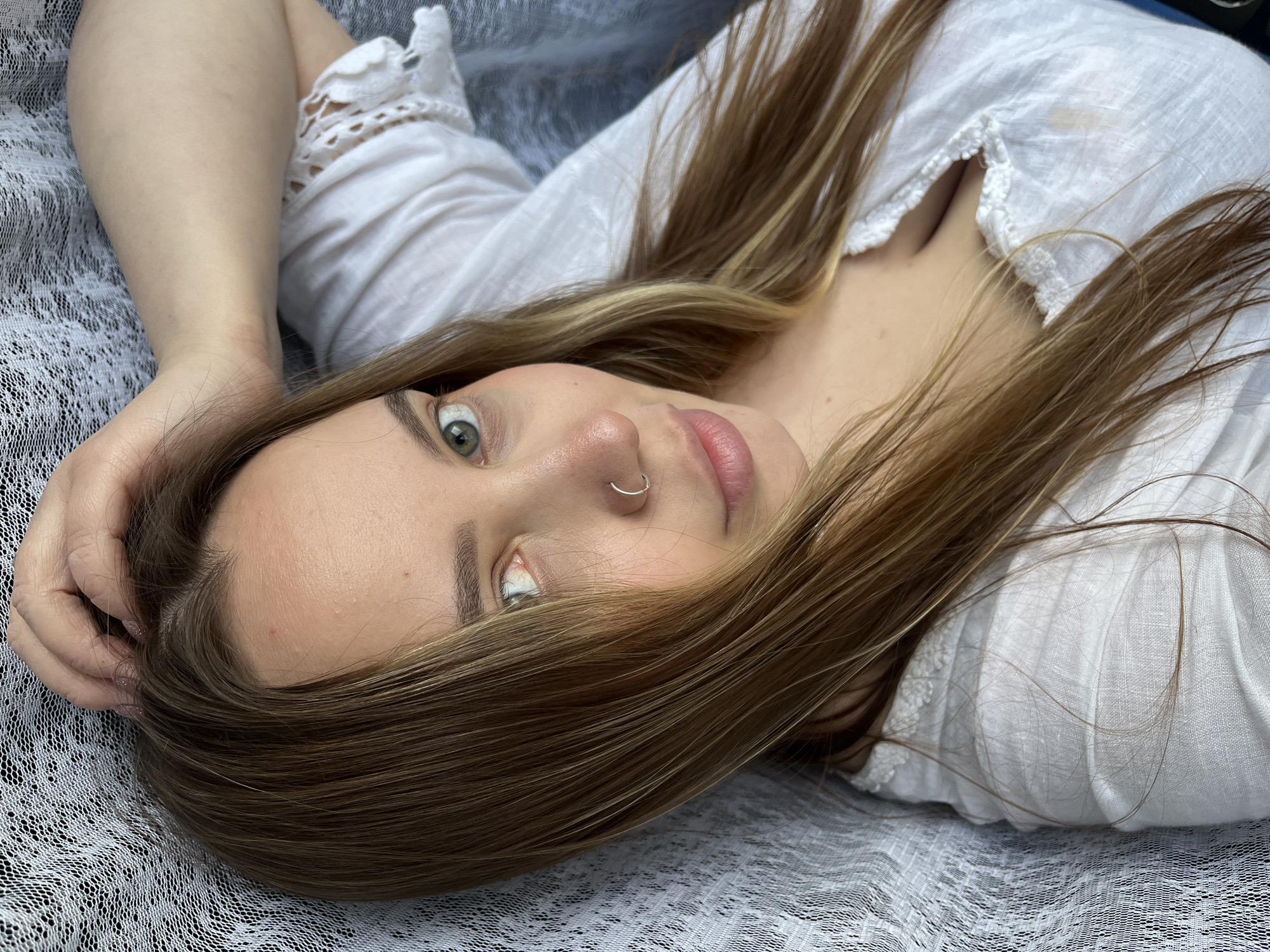 A young woman lies supine on a white, possibly crocheted or lacy, surface. She is wearing a white, long-sleeved blouse with lace detailing. Her hair is long and light brown with blonde highlights. She has fair skin, light-colored eyes, and a silver nose ring. One of her arms is bent and positioned near her head, the hand partially obscuring her forehead. The location is unspecified but reported to be in the city of Novopokrovskaya, Russia. No specific actions or interactions are visible other than the woman lying down.