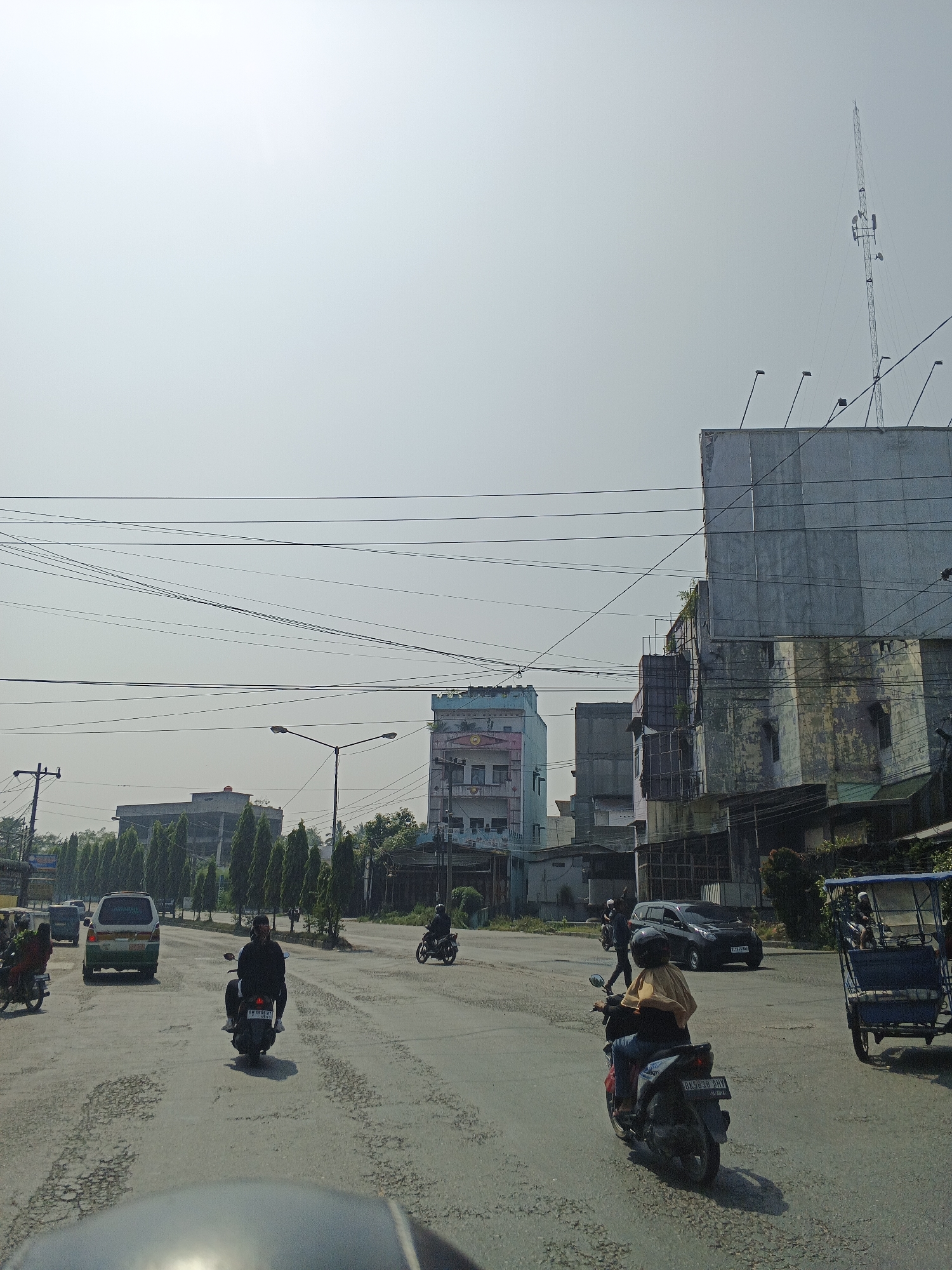 The image depicts a street scene in Tg Morawa A, Indonesia, under a hazy, bright sky. The road surface is heavily deteriorated, with numerous potholes and cracks. Several vehicles are present: a white van is visible on the left, a black car is in the middle of the road, and a blue cart, possibly a rickshaw, is on the right. Multiple individuals are riding motorcycles. One motorcycle in the foreground has two occupants, a rider wearing a black jacket and a passenger wearing a light-colored scarf. Another motorcycle further back has a rider wearing a yellow garment and jeans. Two more motorcycles are seen in the distance. Buildings line the sides of the street, with a notable tall structure painted light blue with pink accents in the mid-ground. Electrical wires crisscross the sky, and a communication tower stands on the right side of the frame. The overall atmosphere suggests a busy urban environment on a clear but possibly polluted day.