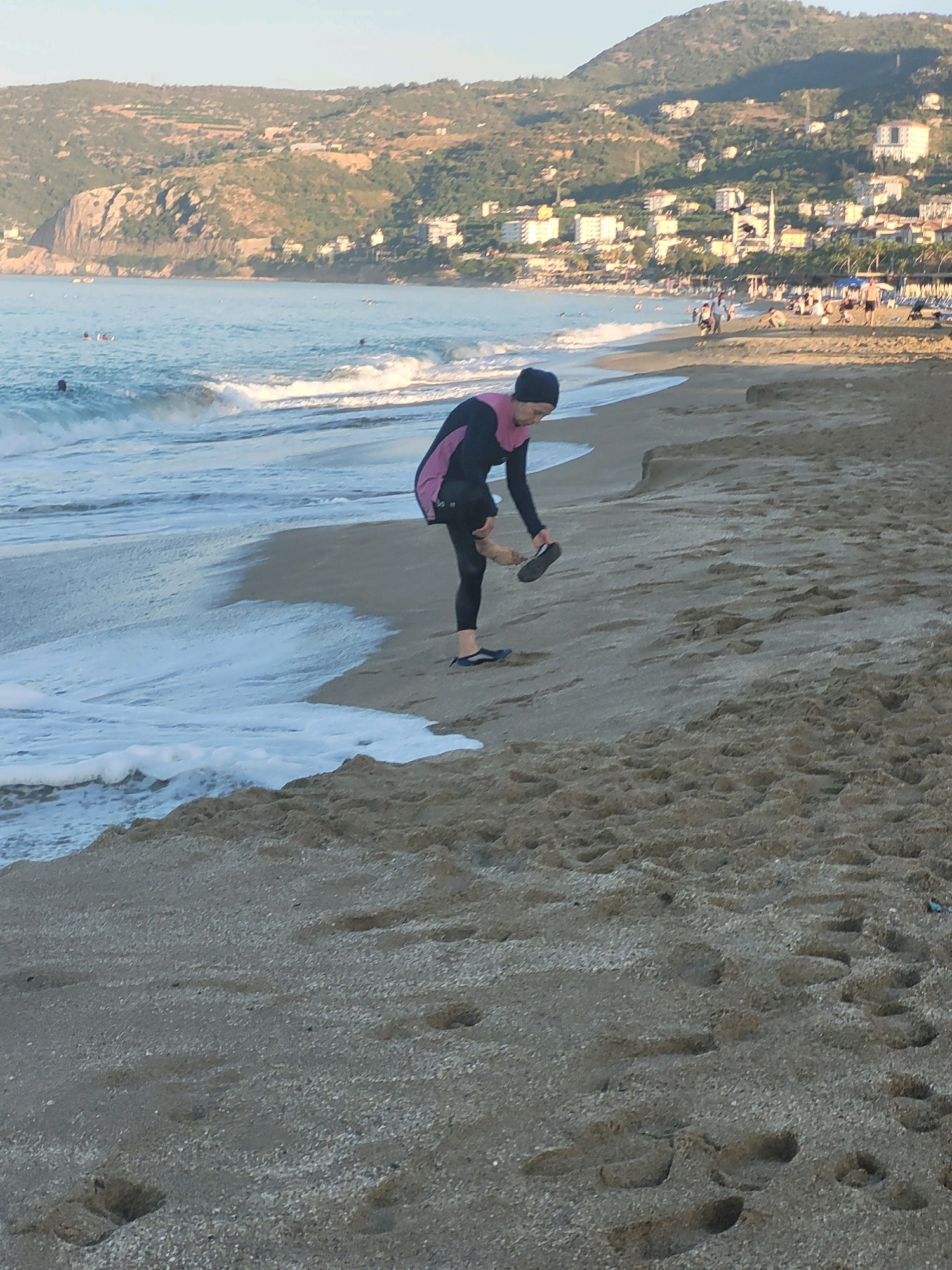 A person wearing a black full-body swimsuit and a dark head covering stands on a sandy beach in Alanya, Turkey. The person is facing right and is bending over, holding a dark sandal in their right hand. Their left foot is bare and positioned on the sand. The water of the sea is to the left of the person, with waves gently lapping at the shore. In the background, there are more people on the beach, some swimming in the sea, and buildings nestled into a hillside that rises behind the coastline. The sky is clear and a pale blue, suggesting either early morning or late afternoon lighting. The sand is a light brown color and has numerous footprints visible.