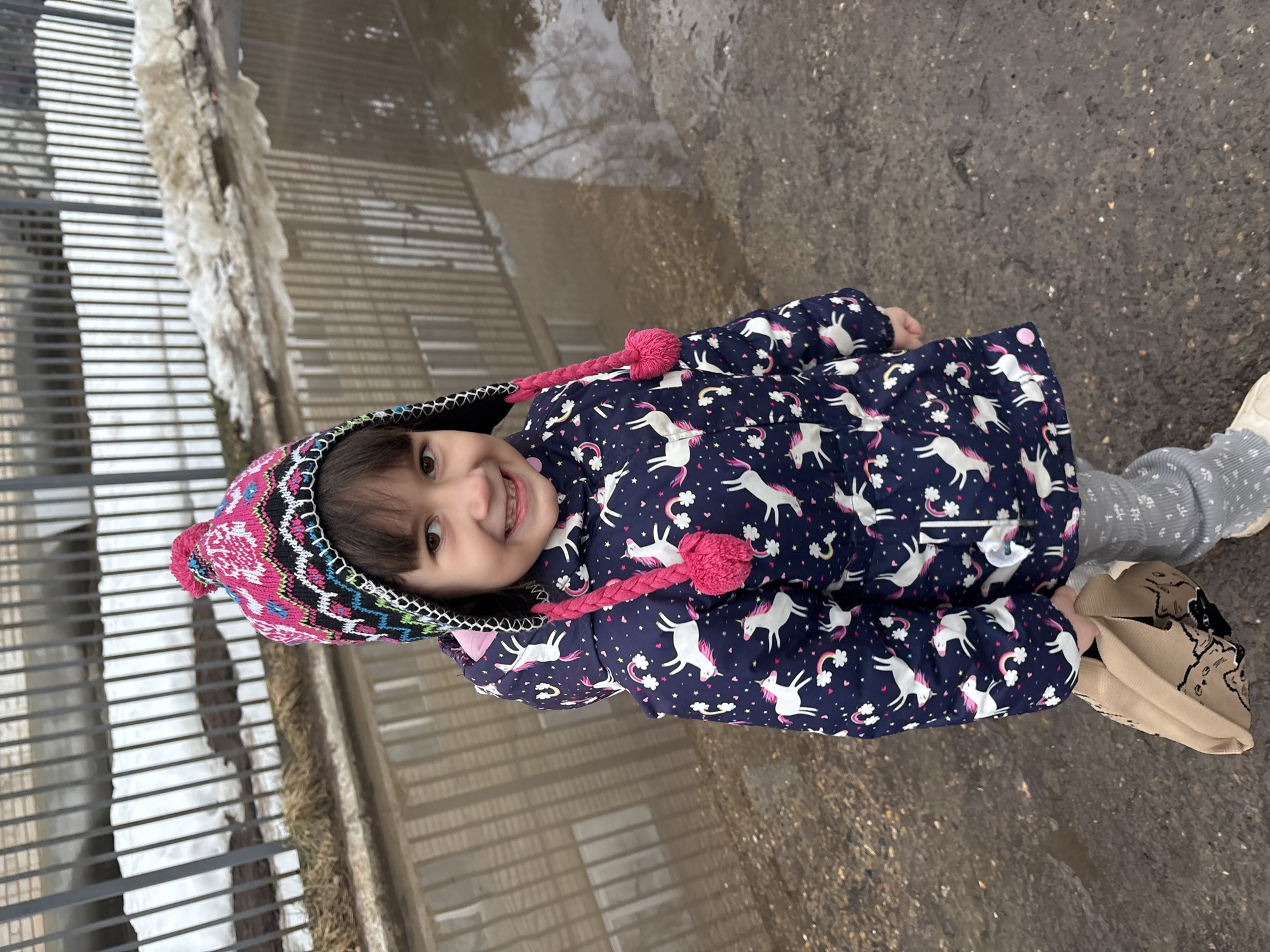 A young girl, estimated to be a toddler or preschooler, with dark hair and a visible partial orthodontic appliance on her upper teeth, stands outdoors, smiling directly at the viewer. She is wearing a multicolored knit hat featuring pink, black, white, and light blue patterns, adorned with a large pink pompom on top and two braided pink ear flaps ending in smaller pompoms. Her attire includes a navy blue padded jacket patterned with white unicorns and small multicolored rainbows. Her lower body is covered by light grey, textured leggings, and light-colored shoes. In her left hand, she holds a crumpled light brown bag with a black outline design. The girl is positioned on dark, wet ground, partially standing in a reflective puddle that covers the surface behind and to her right. In the background, a dark metal fence with vertical bars is visible, behind which remnants of dirty, melting snow and patches of dry, light-colored vegetation are present. The environment appears to be an urban or semi-urban setting during a thaw, located in Chaikovsky, Russia, though the exact coordinates within the city are not discernible from the image.