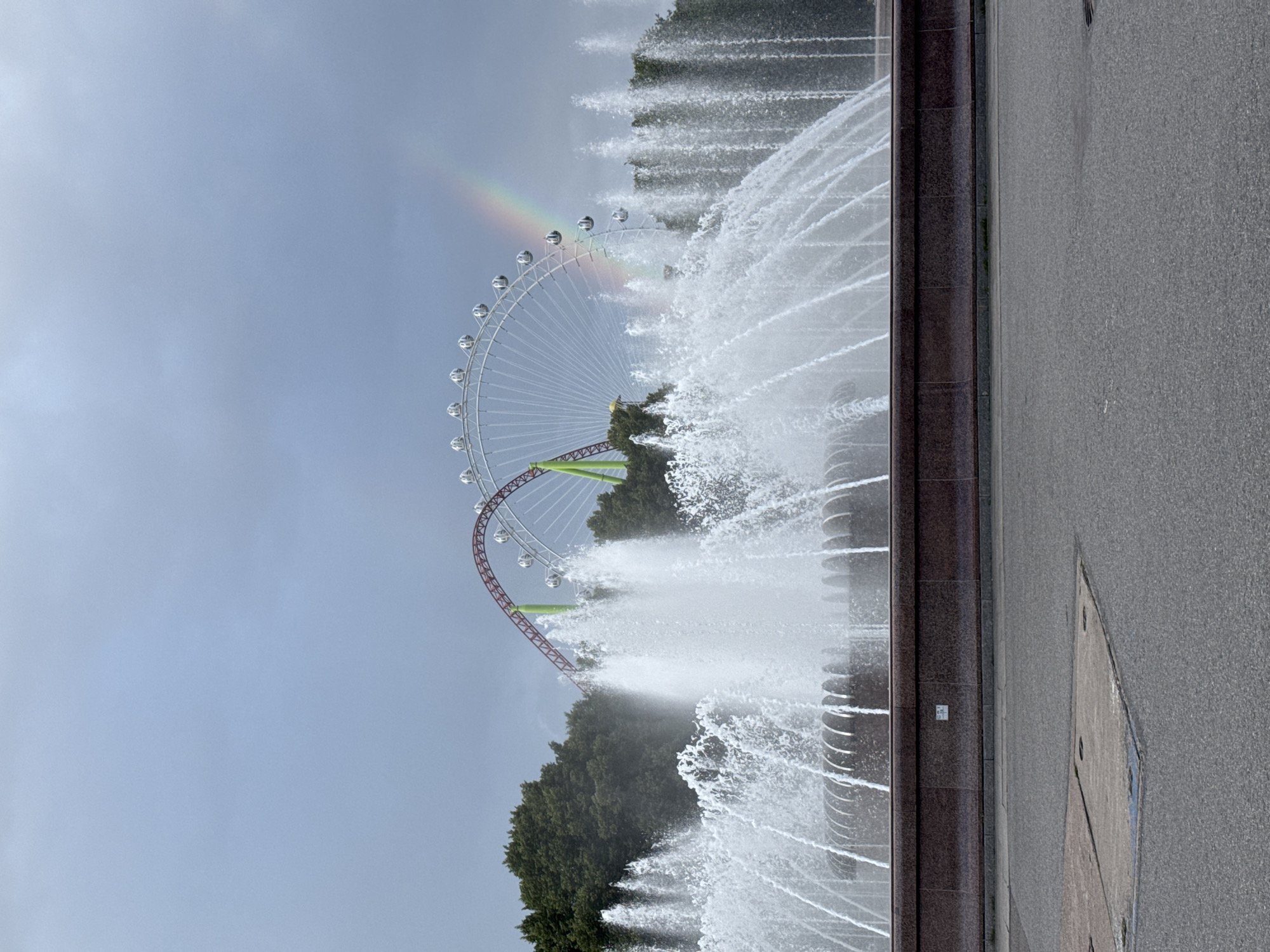 The image shows a large fountain in the foreground, with water jets spraying upwards and outwards. Behind the fountain, there is a Ferris wheel and a roller coaster, partially obscured by trees. A rainbow is visible in the sky above the Ferris wheel. The sky is overcast and a bit cloudy. The scene is likely in an amusement park or a public park with attractions. This specific location is in Saint Petersburg, Russia, and it is the historic park of Dynamo, with the Ferris wheel of the “Dixie Land” amusement park and the water park attractions of the large fountain, located near the Neva river. The fountain is in front of the monument to the Soviet soldiers.