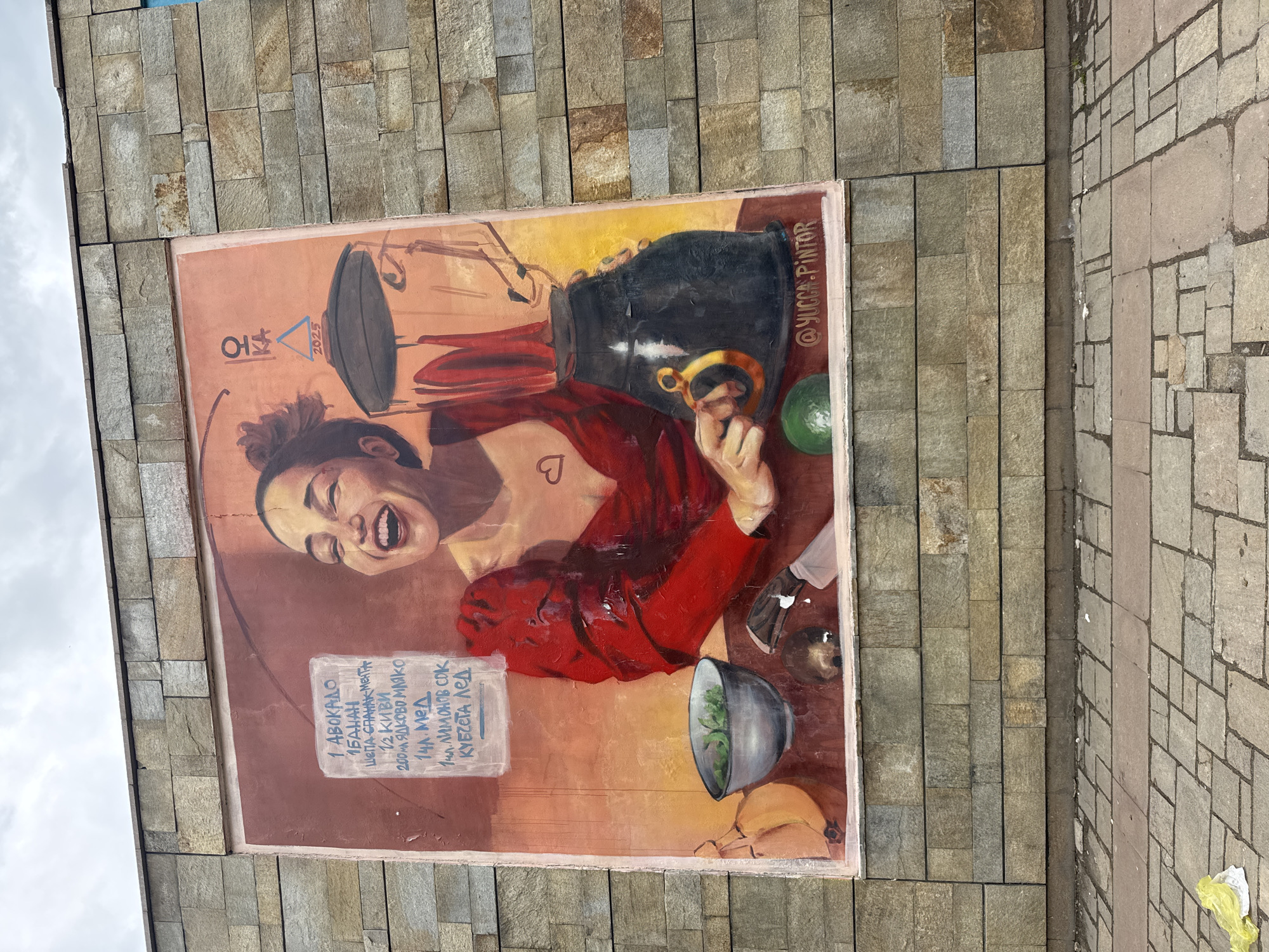 A vibrant mural, located in Sofia, Bulgaria, adorns a wall made of rectangular stone tiles. The artwork features a woman with dark hair tied up, dressed in a red top, laughing heartily with her eyes closed and mouth wide open. A small heart shape is visible on her chest. She is positioned amidst various ingredients and kitchenware, suggesting the preparation of a healthy beverage.
To her right, a large dark pot or blender with a golden handle is prominent. Below her, a grey bowl contains green leafy ingredients, possibly spinach. Other items like an avocado and kiwi are also depicted. A list of ingredients is painted on a white rectangular background to her left, written in Bulgarian: "1 AVOCADO", "1 BANANA", "Handful of SPINACH Mint", "2 KIWI", "200ml NUT MILK", "1 tbsp Honey", "1 tbsp Lemon Juice", and "ICE CUBES", implying a recipe for a smoothie.
The mural is signed with "@YUCCA.PINTOR" on the right side and displays the year "2025" alongside a symbolic triangle in the upper left corner. The wall is adjacent to a paved ground. The visible sky overhead is overcast, indicating a daytime scene. The painting style is lively and expressive, emphasizing the woman's joyous emotion.