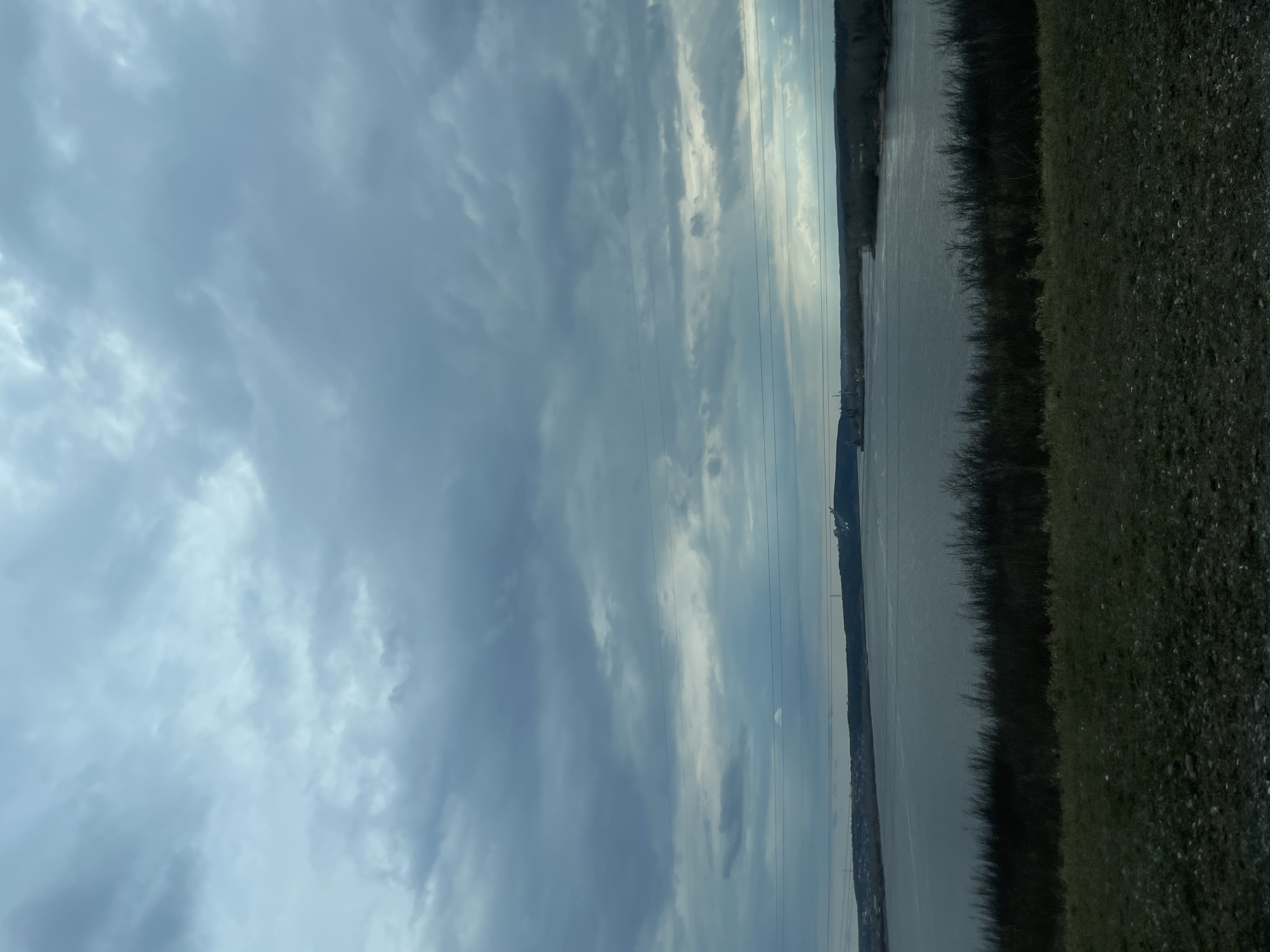 The image depicts a landscape in Chaikovsky, Russia. A body of water, likely the Kama River, stretches across the middle ground of the frame. The river's surface appears somewhat reflective. Along the right edge is a bank covered with vegetation, including grass and possibly some shrubs or trees. The left portion of the image is dominated by a cloudy sky. Overhead power lines are visible. In the distance, along the horizon, there are indications of urban development, including buildings. No people are visible.