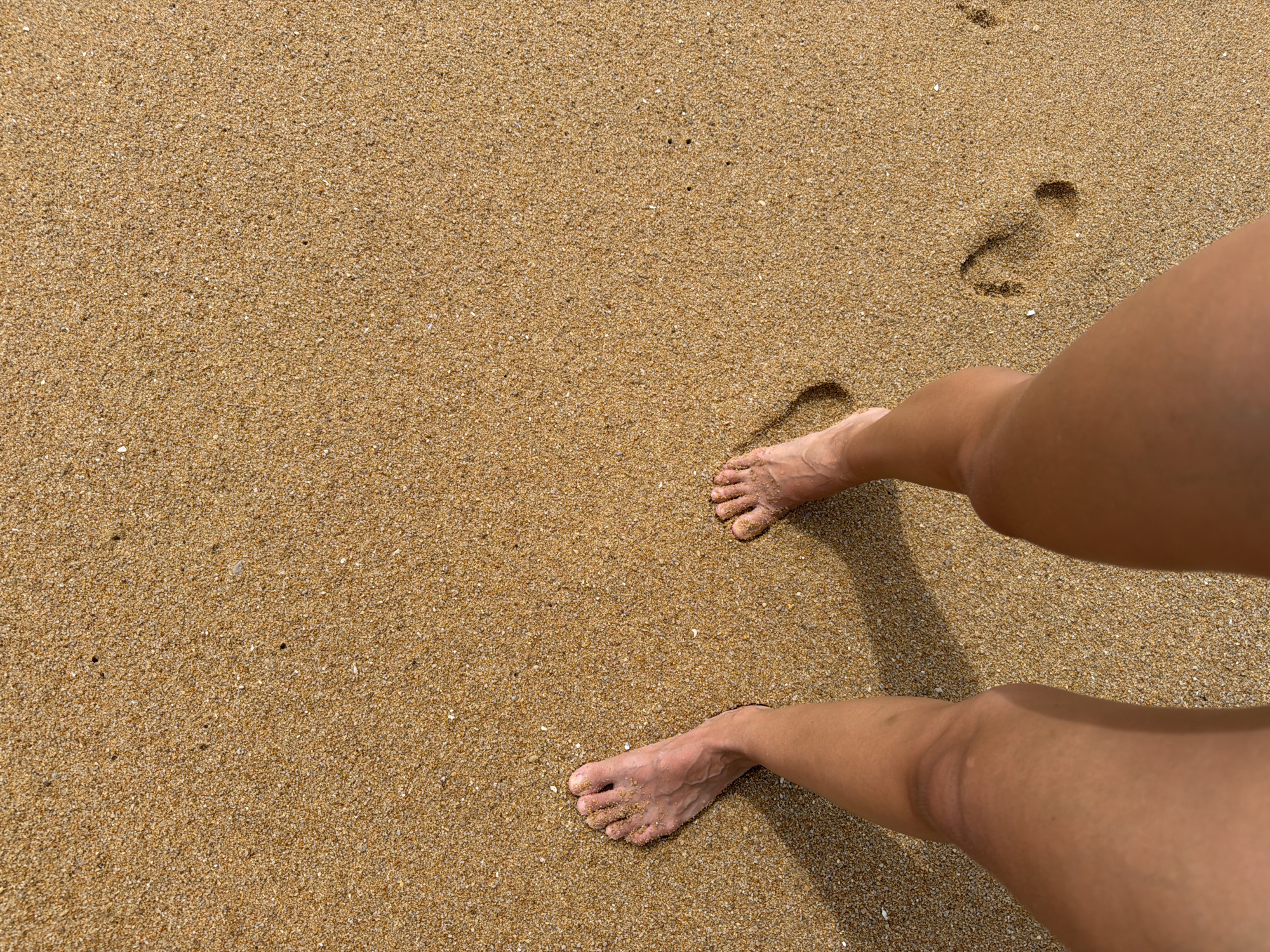 The image shows a person's legs and feet walking on a sandy beach. The sand is a light brown color with small flecks of darker material. There are several footprints visible in the sand, indicating that the person has been walking for some time. The lighting suggests it is daytime, with shadows cast by the person's legs. The scene is likely a beach in Mai Khao, Thailand.