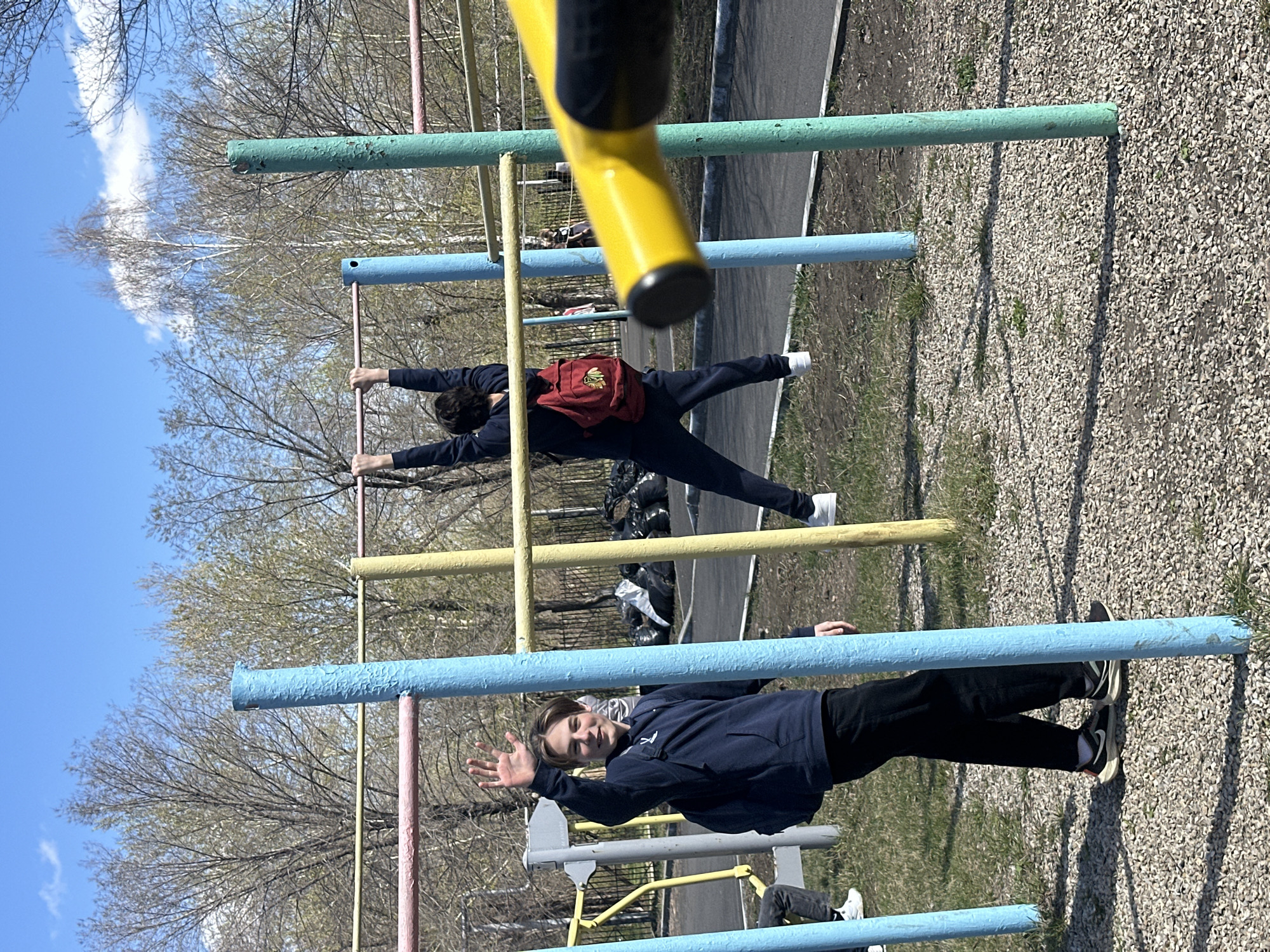 A daytime outdoor scene captures two young men interacting with colorful workout or playground equipment under a clear blue sky with sparse clouds. The image is taken at a slight upward angle, looking through a foreground yellow bar.
In the center-right, one young man, wearing a dark long-sleeved top and dark pants with white sneakers, is performing an exercise or stretch. He holds onto a yellow horizontal bar with both hands, extending his arms and one leg horizontally, while wearing a red backpack with an indistinct emblem.
To the lower-left, another young man, dressed in a dark blue hoodie, black pants, and black and white sneakers, stands near a light blue horizontal bar. He is looking towards the viewer with a friendly expression, his mouth slightly open in a smile, and one hand raised in a wave.
The workout equipment features brightly painted metal bars and poles in yellow, light blue, green, and pinkish-red. The ground consists of light-colored gravel or pebbles mixed with patches of green grass. In the background, deciduous trees with mostly bare branches indicate early spring or late autumn. A paved path or road is visible further back, along with some dark plastic bags. Long shadows suggest a sunny day, likely mid-morning or late afternoon. No visible text is clearly legible.